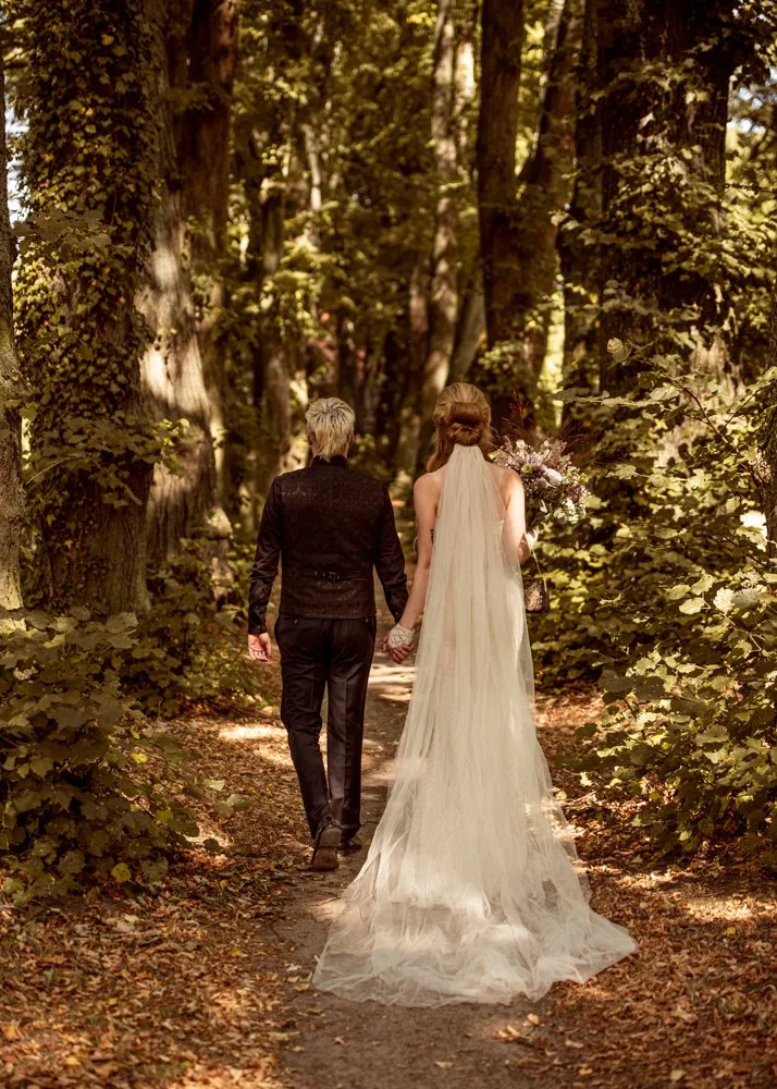 A newlywed couple walking hand-in-hand on a wooded forest trail, with the bride wearing a long white wedding dress and veil, and the groom in a black suit.