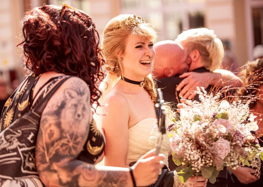 People celebrating at a wedding, with a woman in a strapless dress holding a bouquet of flowers, smiling and wearing a tiara, and others hugging in the background.