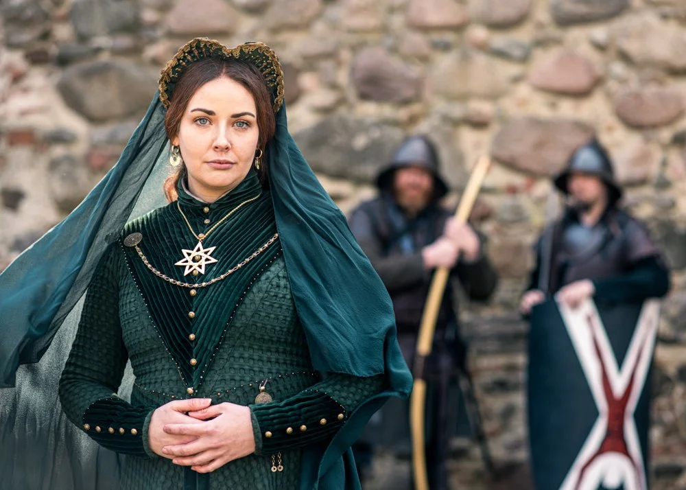 A woman dressed in medieval or fantasy-style clothing with a dark green velvet dress and a headpiece, standing in front of a stone wall. Two men dressed as soldiers or knights, holding weapons and shield, are blurred in the background.