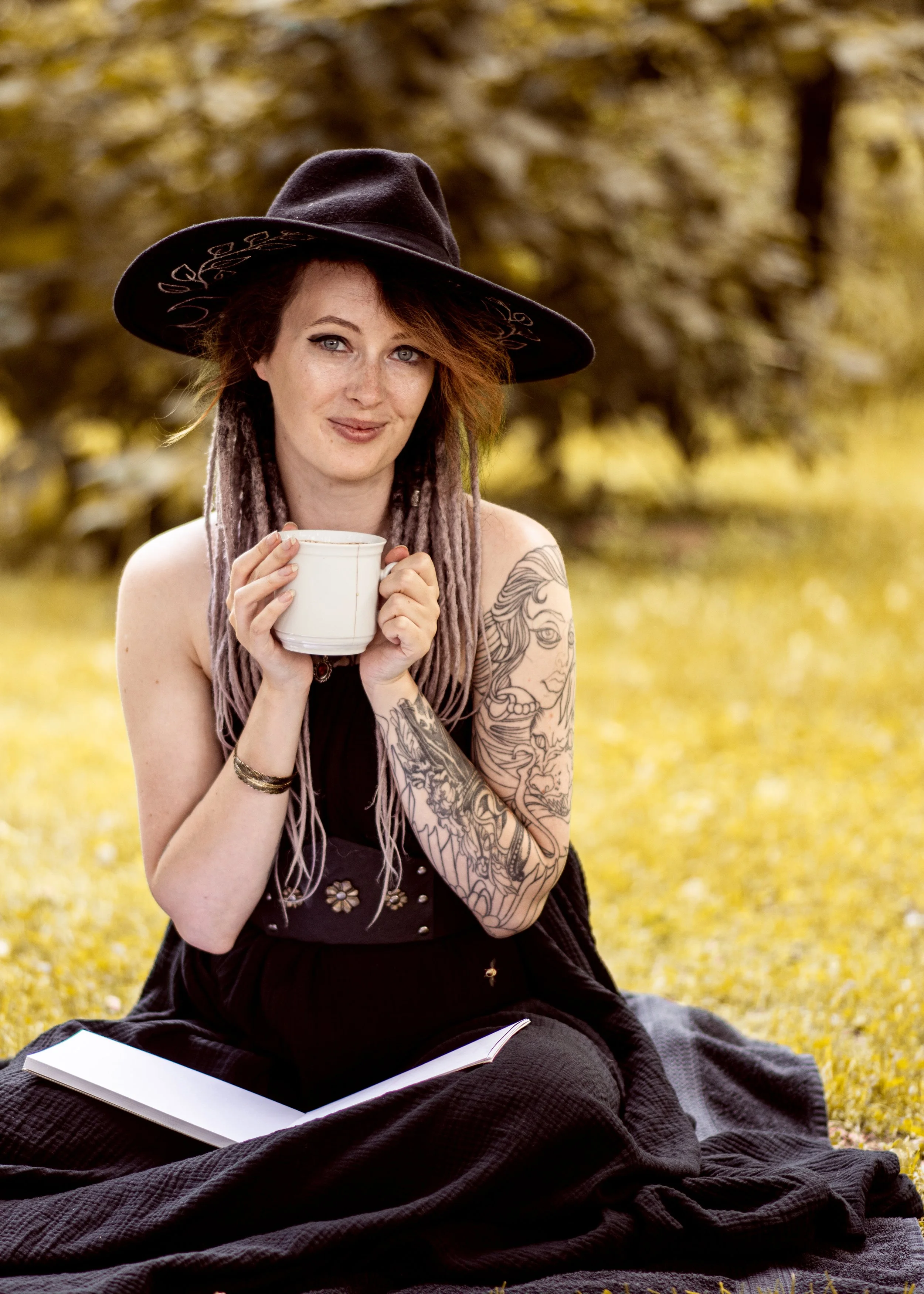 A woman with tattoos on her arm, wearing a large black hat, sitting outdoors on a blanket, holding a white mug, and smiling, with a book open in her lap, in a park during autumn.