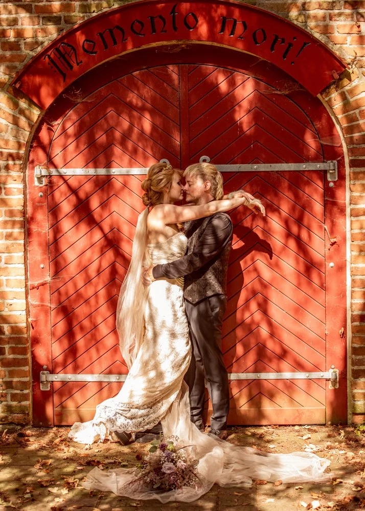 A couple in wedding attire sharing a romantic kiss in front of a red rustic barn door with a sign above that reads 'Memento Mori.' The bride is wearing a white strapless wedding gown, and the groom is dressed in a dark suit. There are fallen leaves on the ground and a bouquet of flowers lying in front of them.
