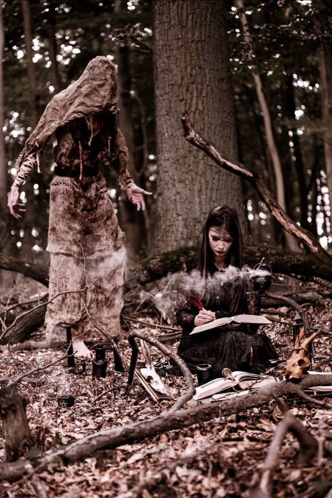 A person with long hair, dressed in dark clothing, sits on the forest ground writing in a book with a red feather quill, amidst fallen branches and trees, with a large tree in the background. Another person dressed in a large, rough, brown costume resembling a tree or creature stands nearby in the wooded area.