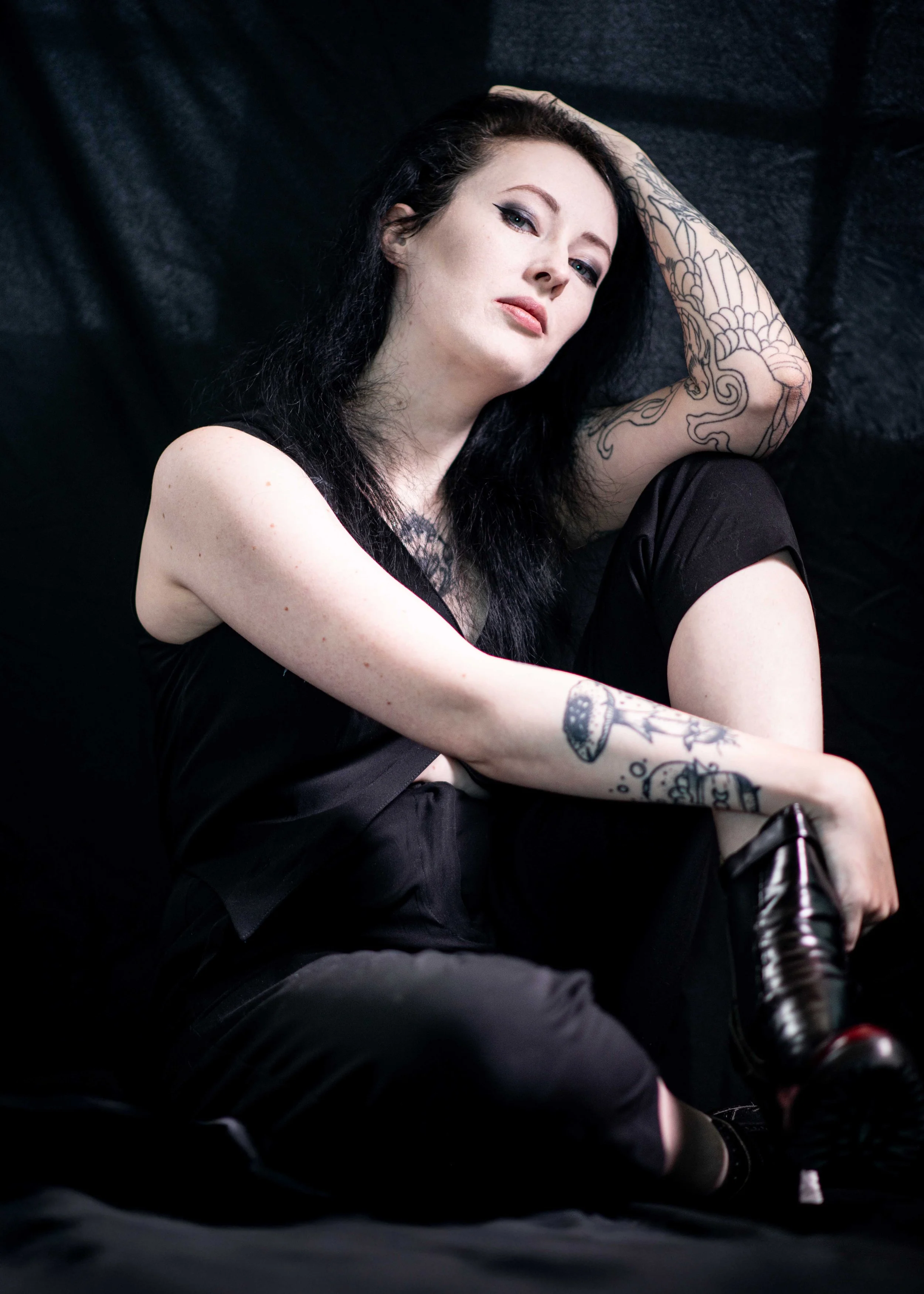 A woman with black hair and tattoos on her arms posing against a black background. She is sitting with her right knee bent and her left arm resting on her head. She wears black clothing and is looking at the camera.