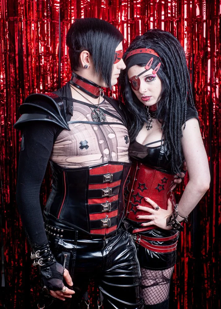 A couple dressed in gothic fashion with red and black outfits and accessories, standing close together against a red metallic streamers backdrop.