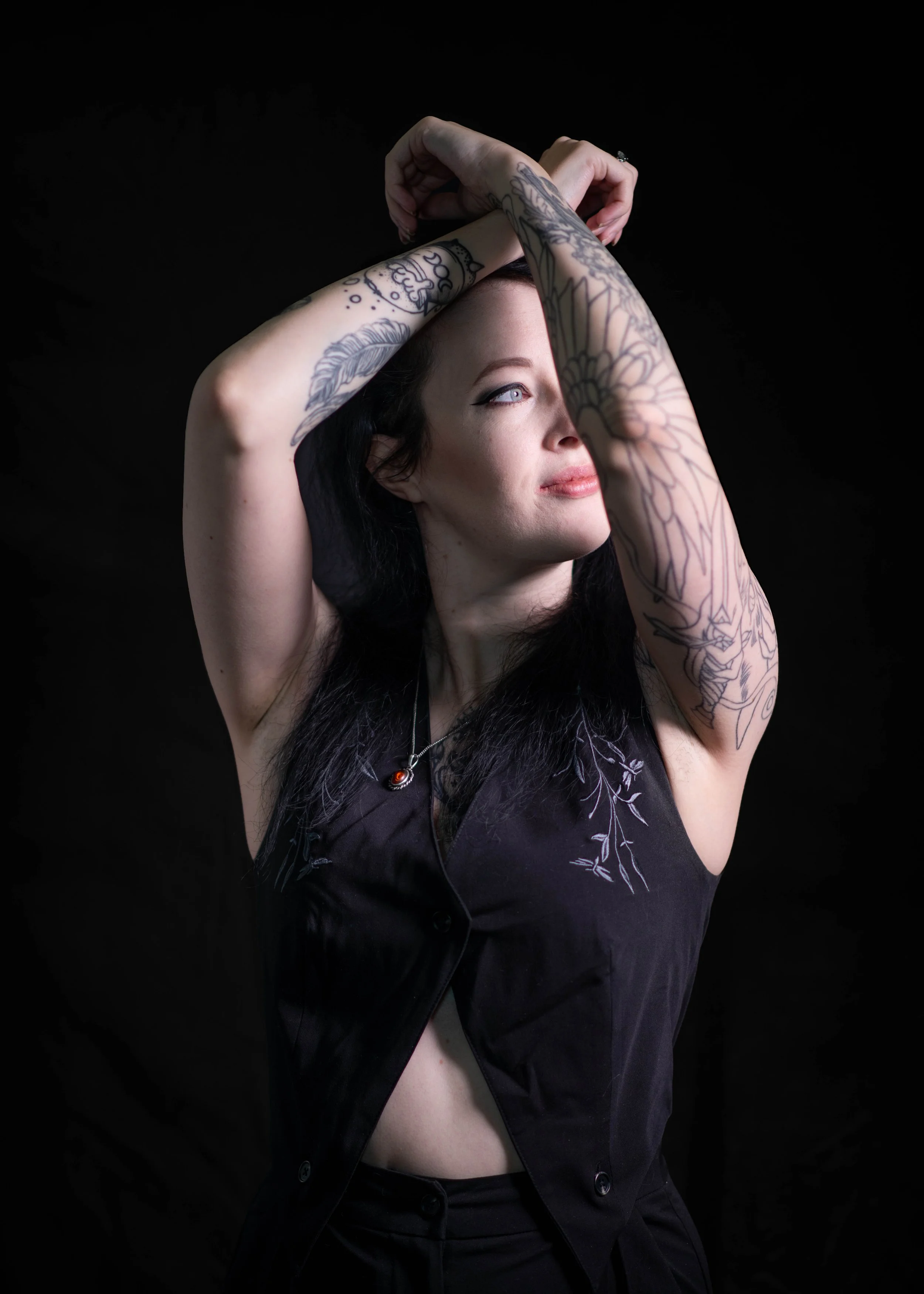 A woman with black hair and tattoos on her arms, wearing a black sleeveless top, looking to the side with her arms crossed above her head against a black background.