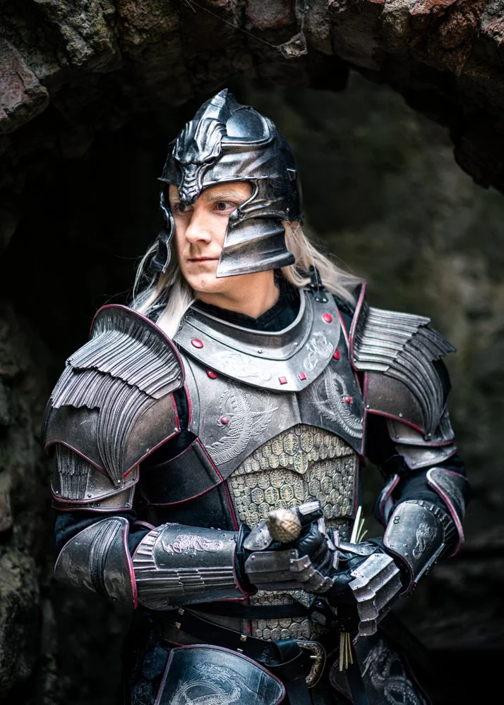 Person dressed as a medieval knight wearing black and silver armor and a helmet, standing near a rocky cave entrance.