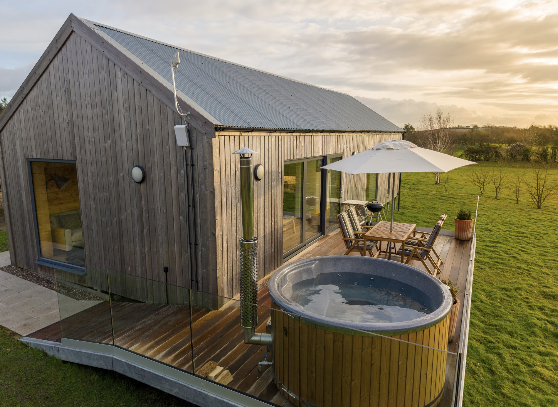 A modern wooden lodge with a large deck, wood fired hot tub, patio furniture with parasol, and a scenic green landscape at sunset.