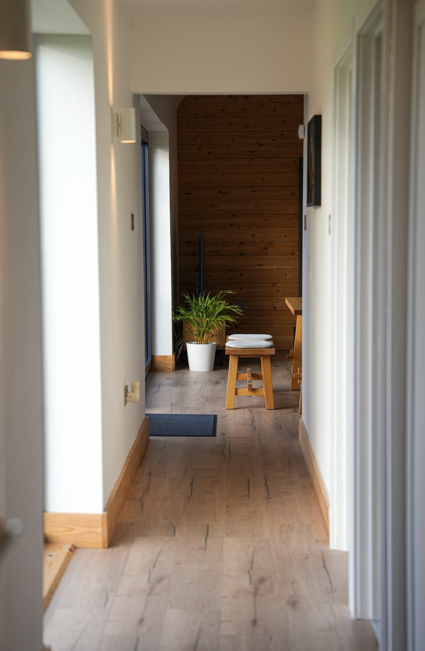 View of a hallway with white walls, wooden floor, and baseboards, leading to a small dining nook with a wooden bench and table, a potted plant, and a wooden paneled wall.