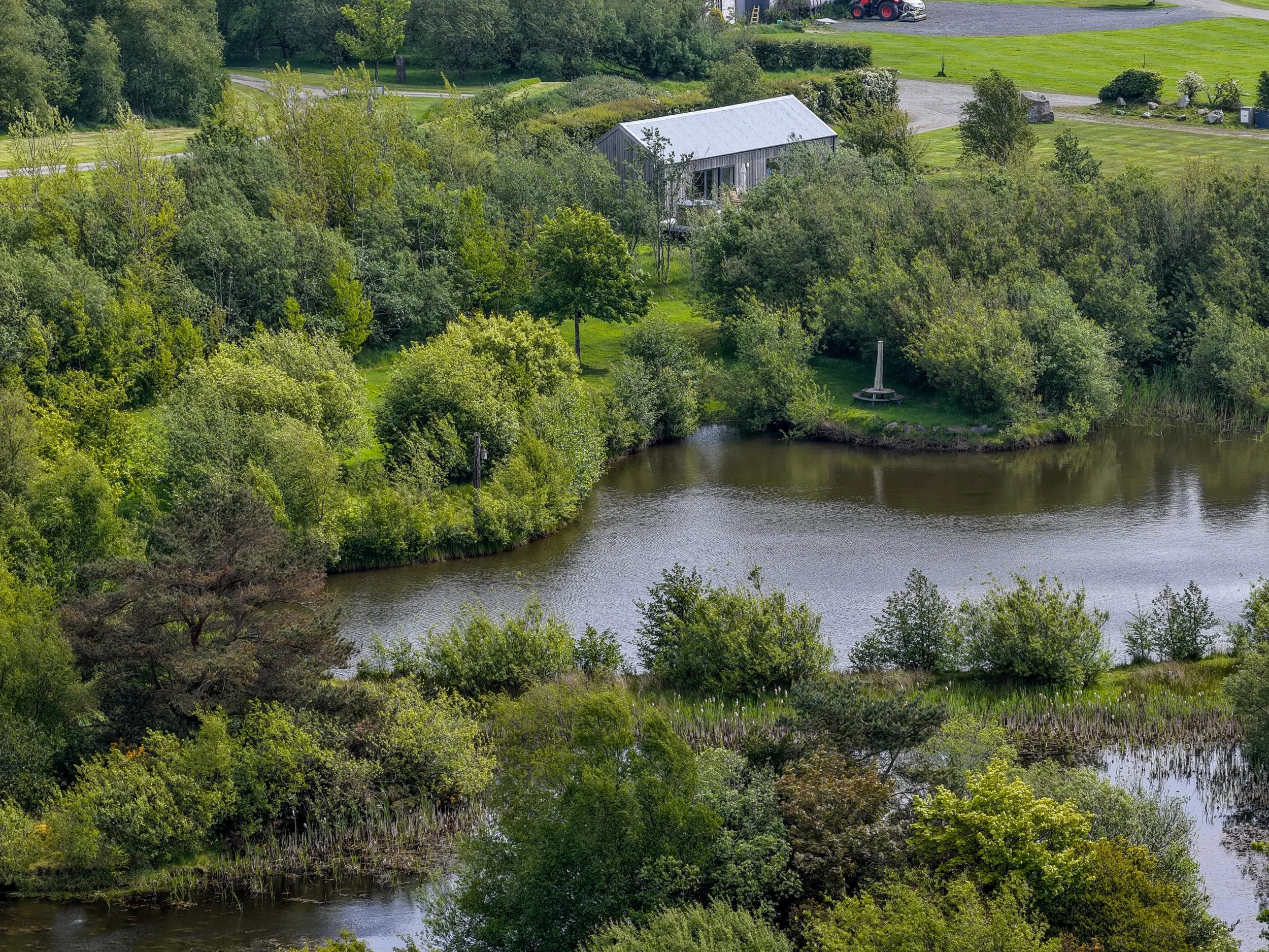 Aerial view of a landscape featuring a body of water, lush green trees, and a gray metal barn surrounded by grass and some structures in the background.
