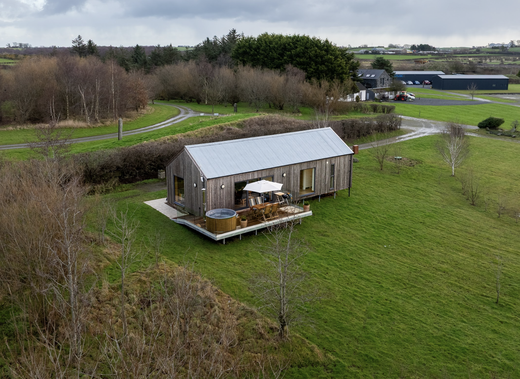 An aerial view of a modern wooden lodge with a metal roof, situated on a grassy area with trees and a deck with outdoor furniture and a hot tub, surrounded by open landscape with a winding driveway.