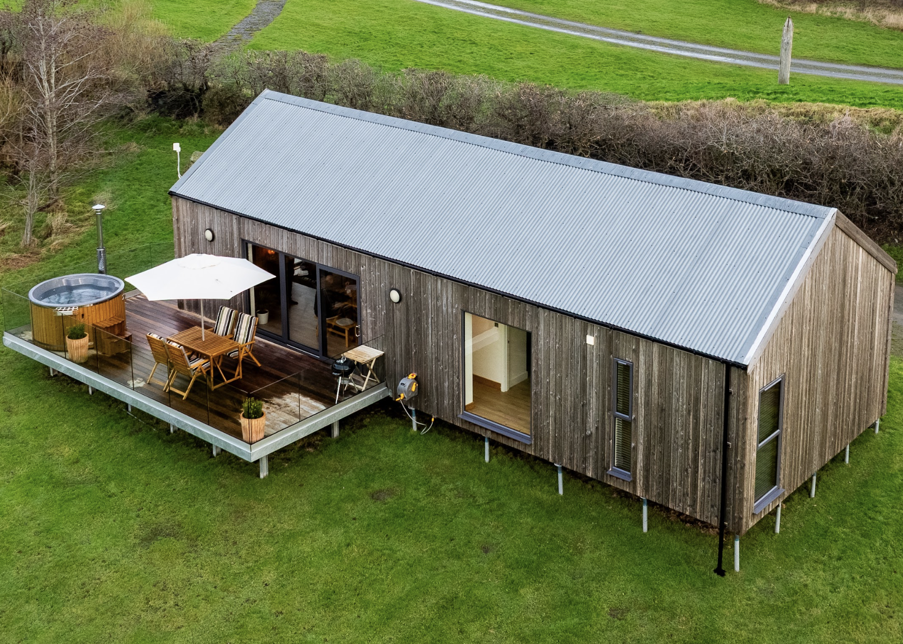 A wooden lodge with a metal roof, an outdoor deck with patio furniture, a hot tub, and a parasol, surrounded by green grass and a wooded area.