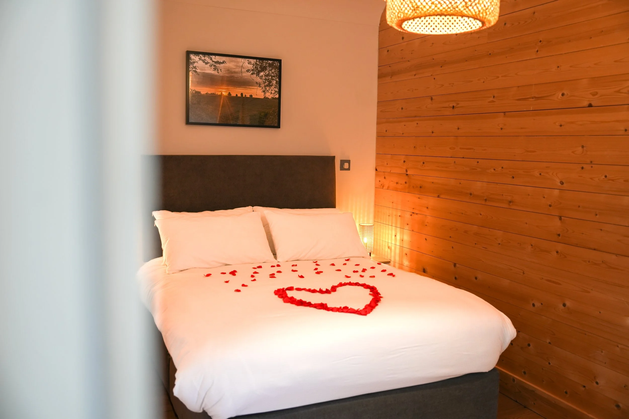 A cozy bedroom with a wooden accent wall, white bed linens, and a heart-shaped rose petal arrangement on the bed. There is a framed sunset landscape picture above the bed and a bedside lamp emitting warm light.