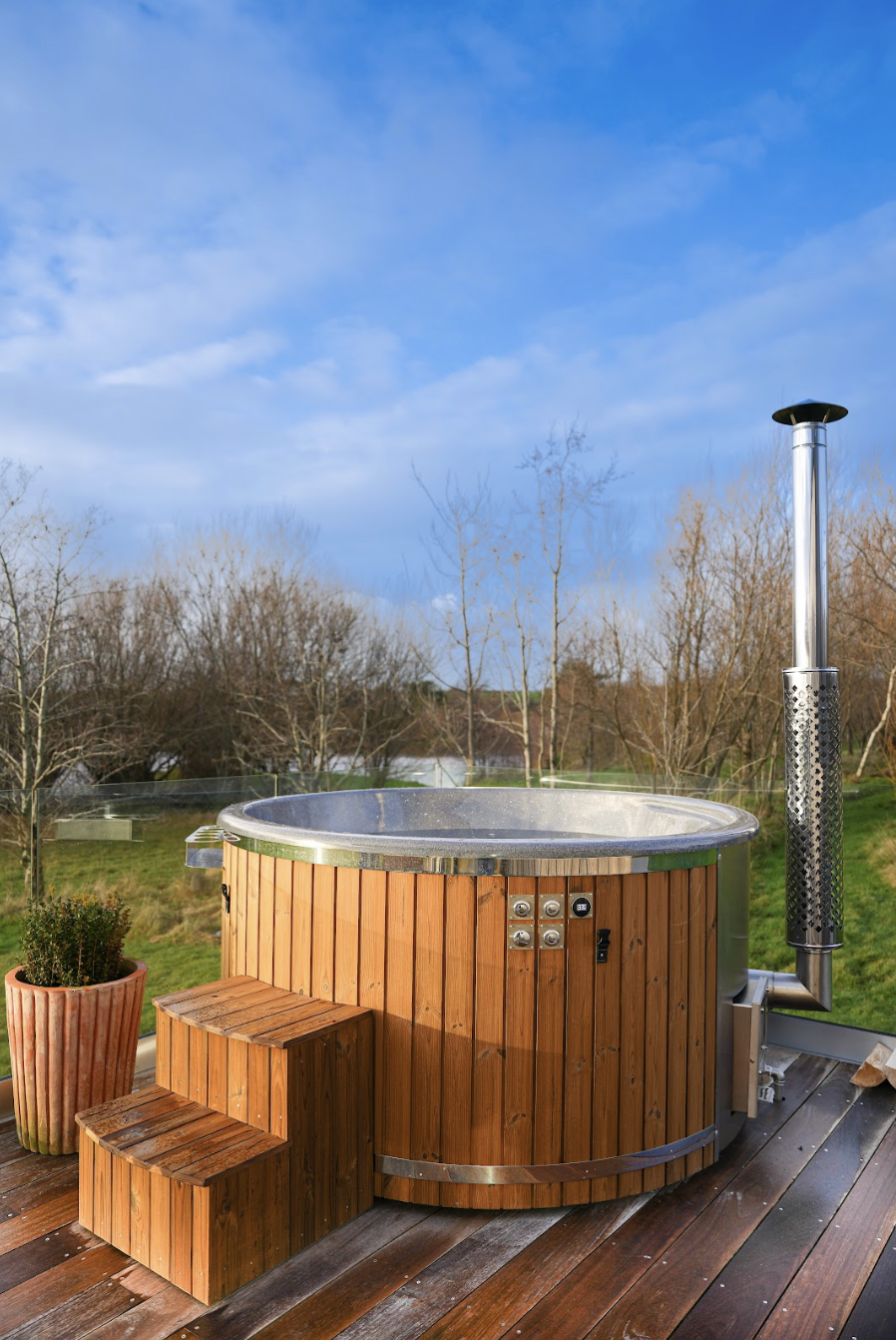 Wood fired hot tub with steps on a private wooden deck overlooking scenic countryside and lake views on a bright blue sky day.
