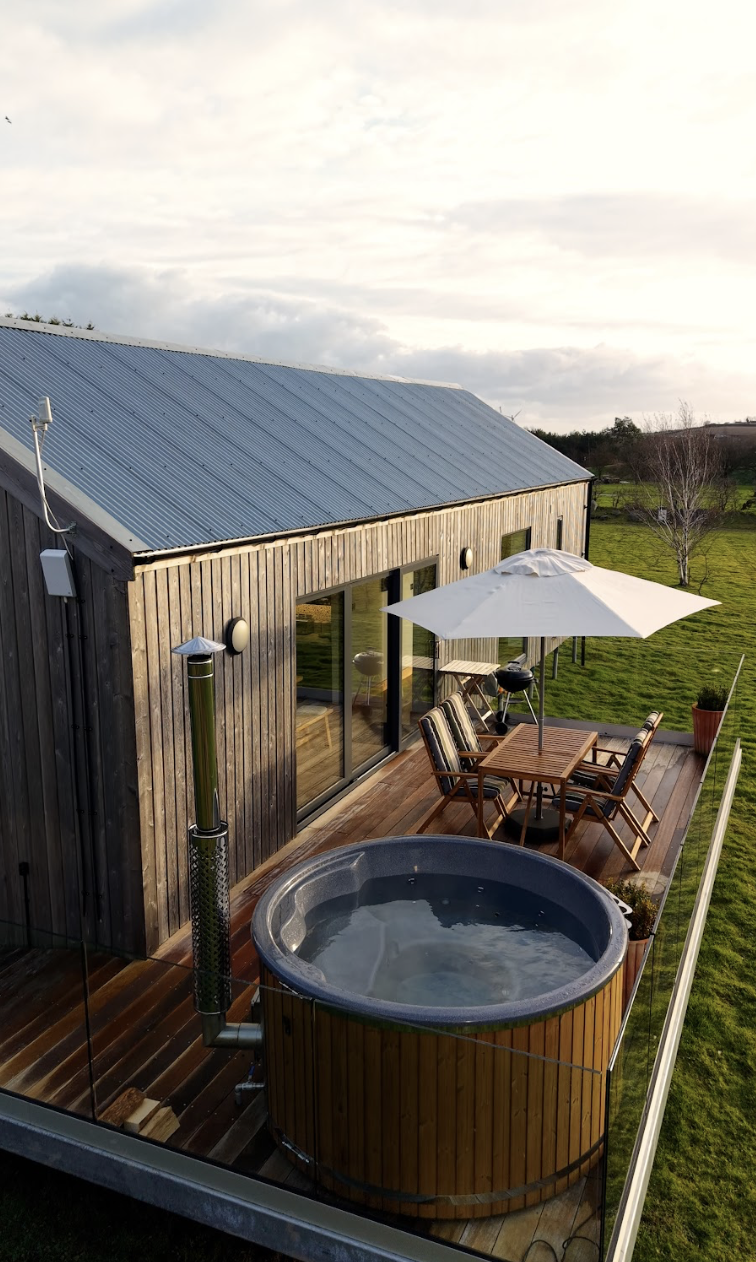 A wooden outdoor patio with a wood fired hot tub, a table with chairs and a parasol, and a grassy yard nearby, attached to a modern lodge with sliding glass doors and a metal roof.