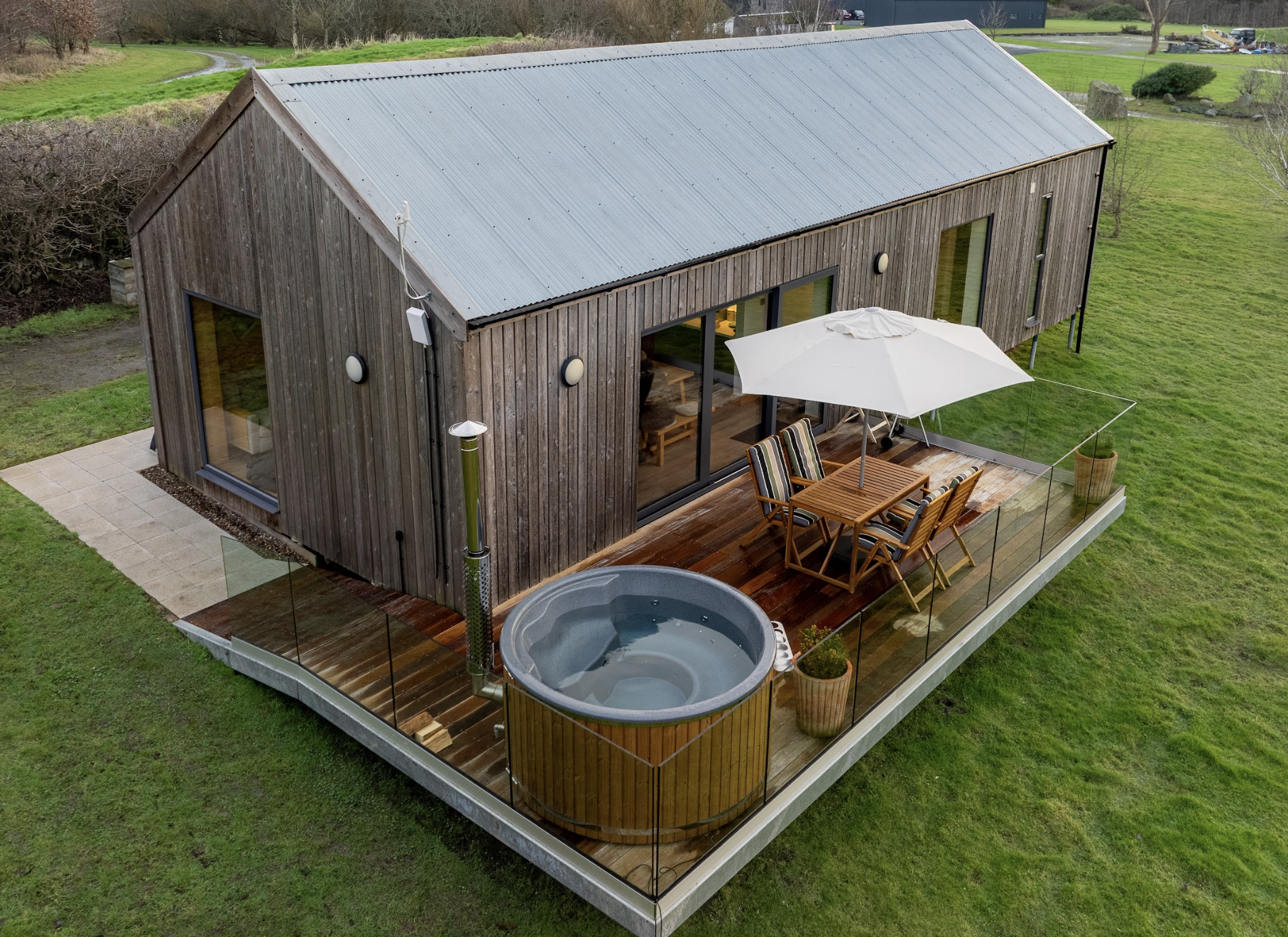 A modern wooden lodge with a metal roof, featuring a large private deck with outdoor furniture, parasol, a wood fired hot tub, and surrounded by a green grassy landscape.