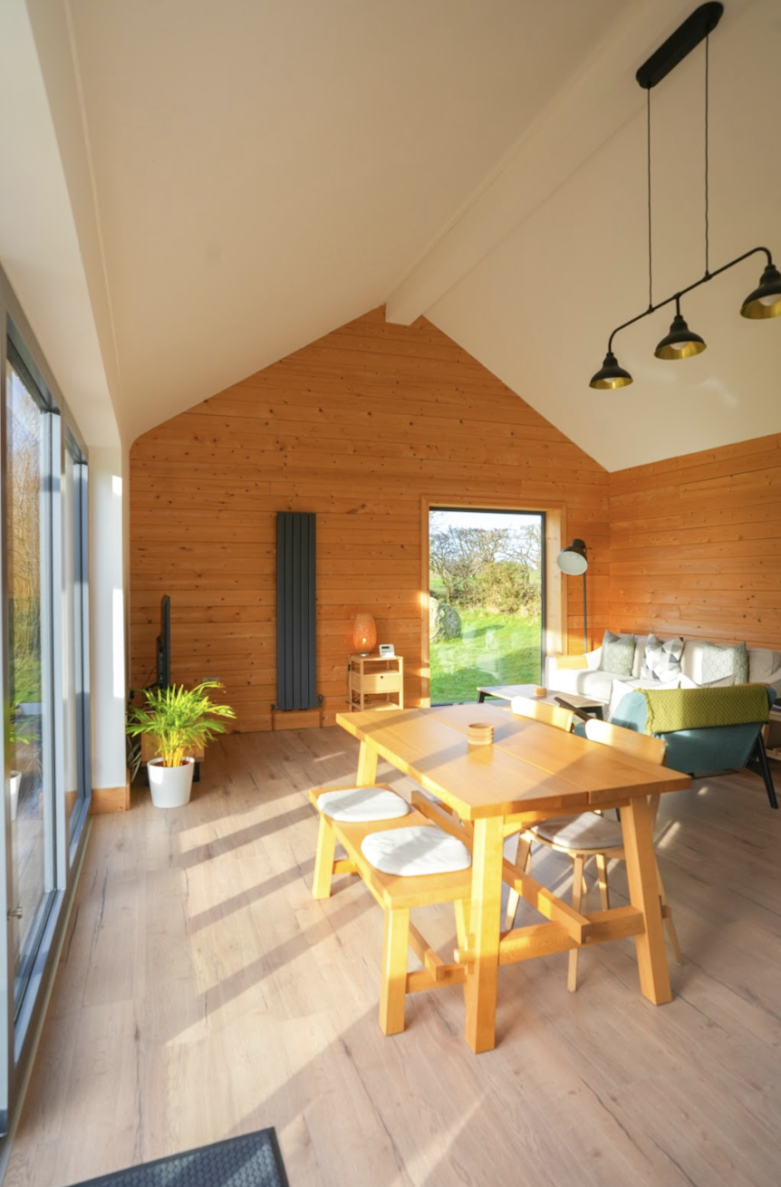 Sunlit living room with wooden walls, dining table and chairs, couch, potted plant, and large windows showing a green outdoor landscape.
