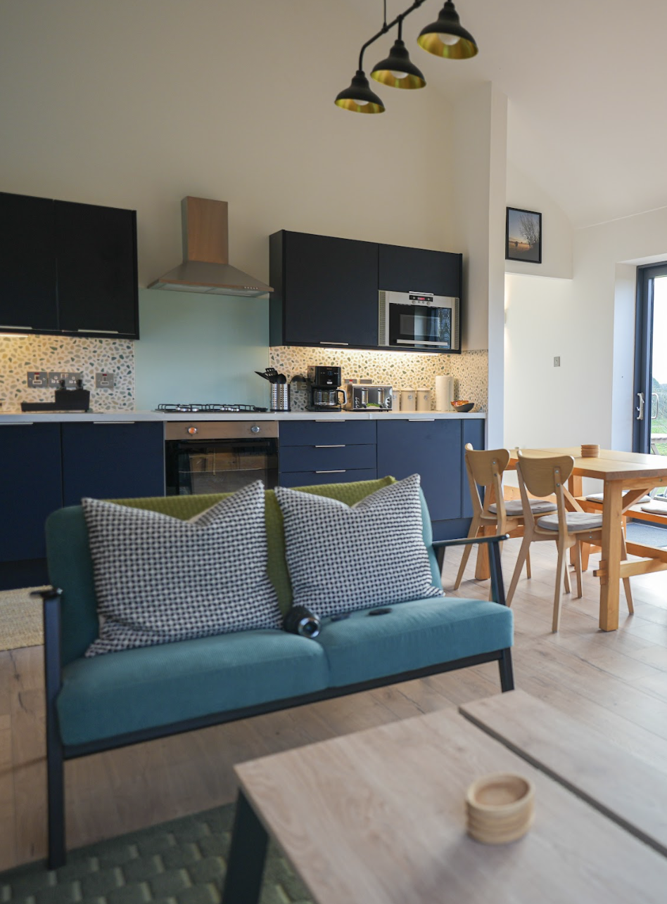 Open kitchen and living room with blue cabinets, a plush couch with cushions, wooden dining table with chairs, and large window/door leading to private decking outside.