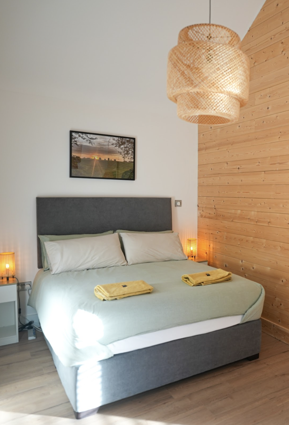 Modern bedroom with a gray upholstered bed, two nightstands with small lamps, yellow towels on the bed, a framed landscape photo above the headboard, and a woven pendant light hanging from the ceiling.