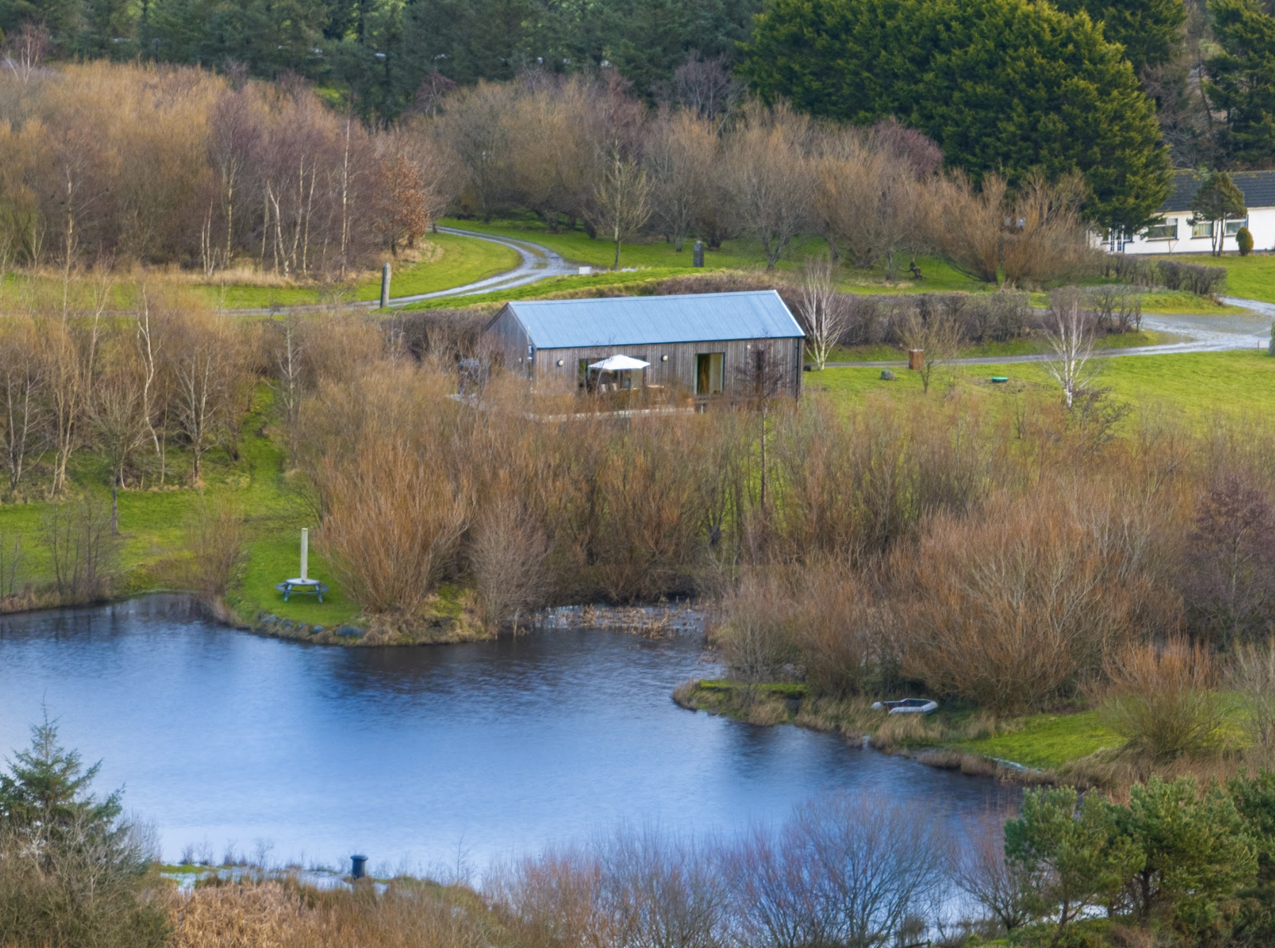 A scenic landscape with a pond in the foreground, a private lodge with a metal roof, and winding roads amidst trees and grassy areas.