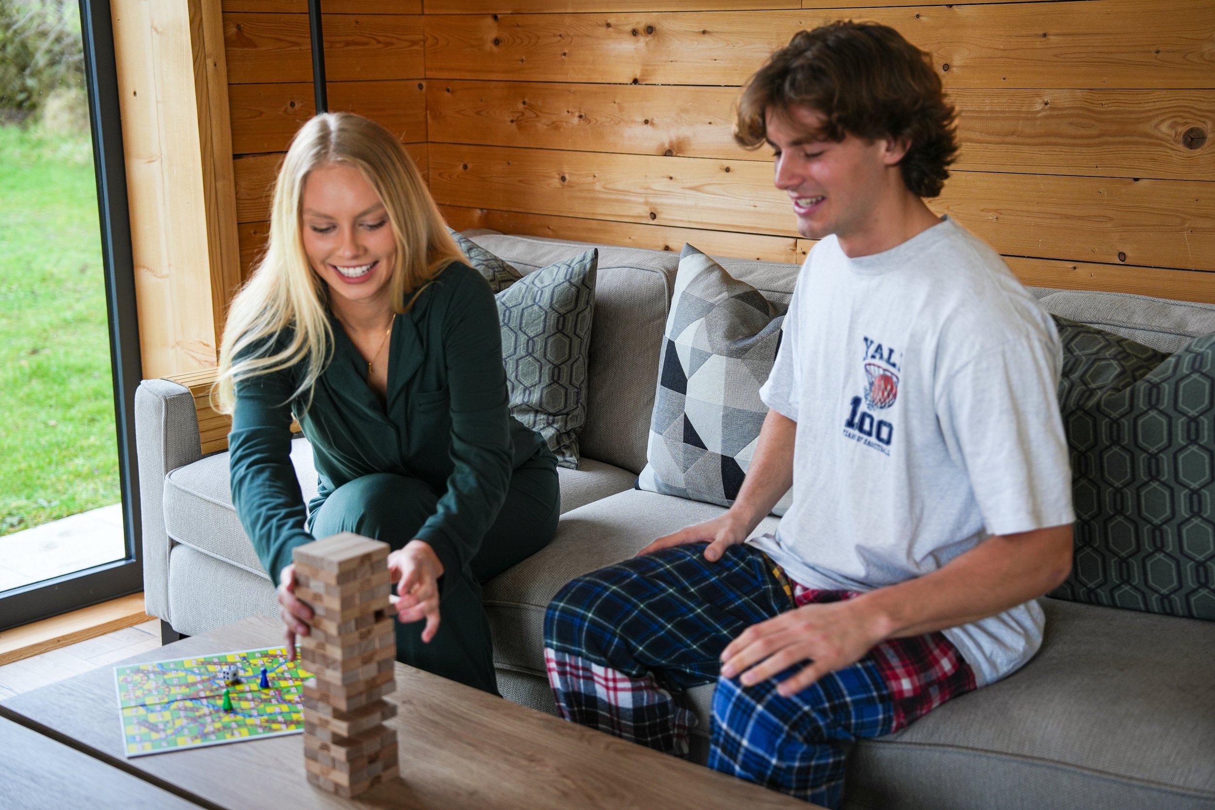 A young woman and a young man are playing Jenga and board games together in a cozy living room with wooden walls and large window, sitting on a gray sofa.