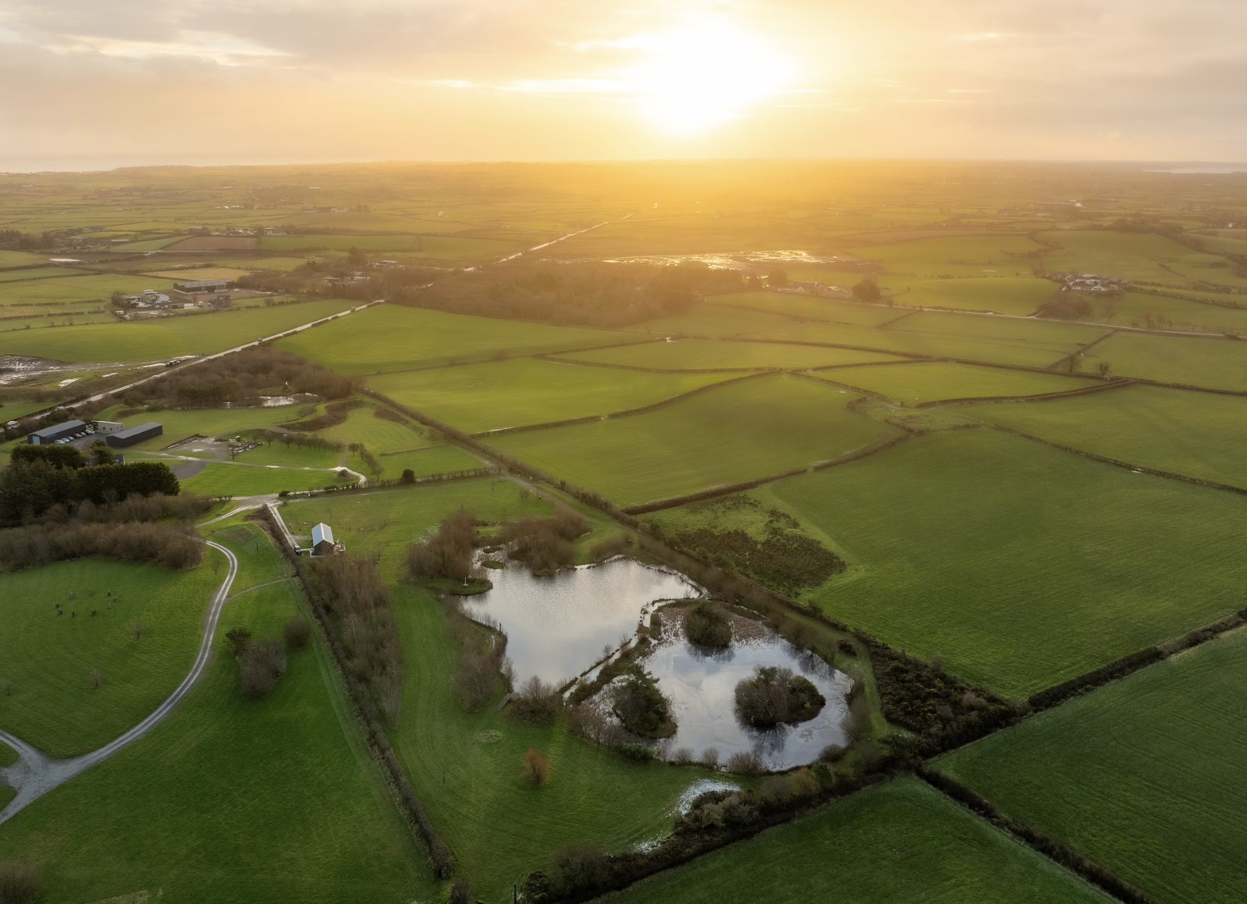An aerial view of lush green farmland with scattered ponds and a private lodge during sunset.