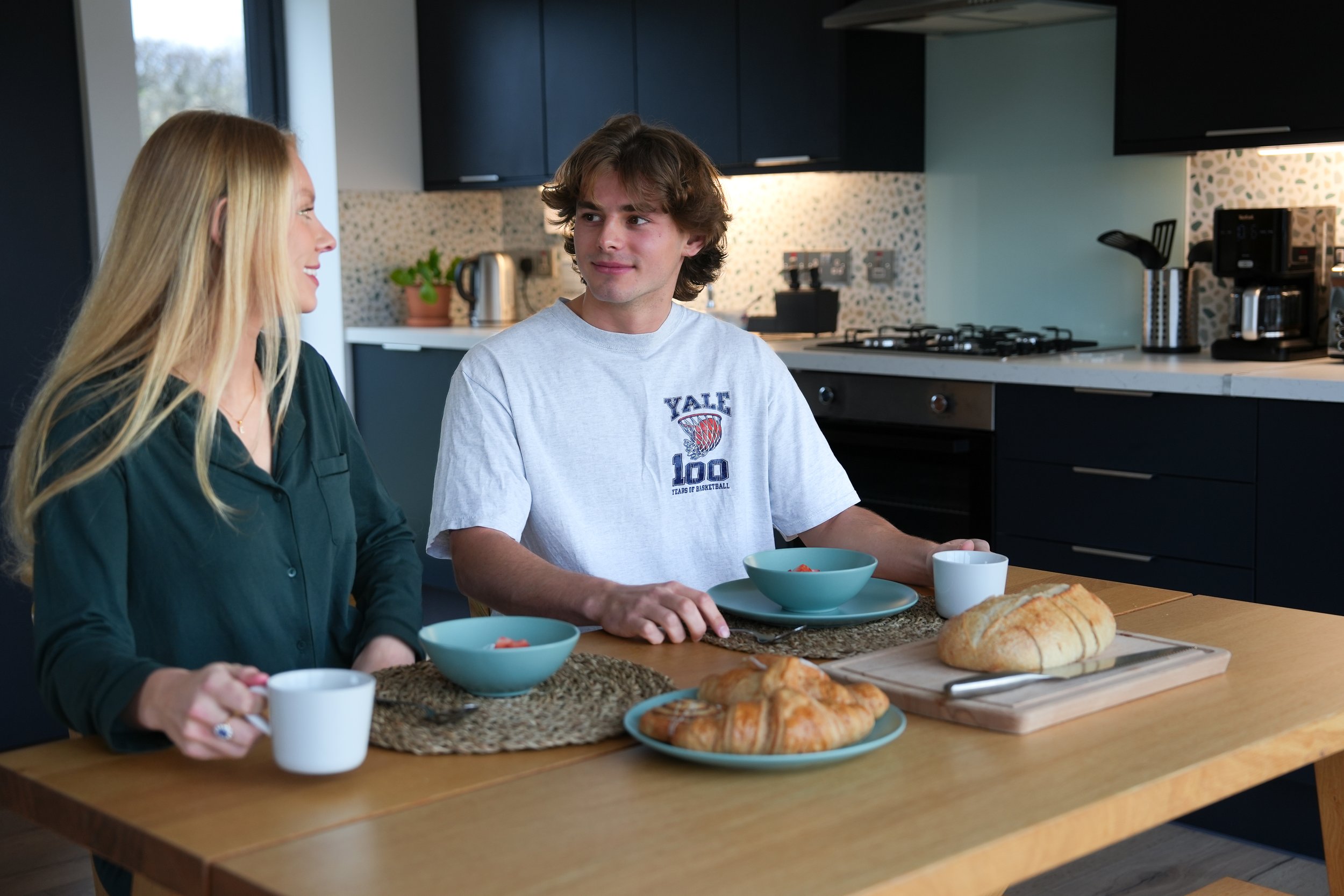 A young man and woman sitting at a kitchen table with breakfast items, including bread, cereal, and cups, engaged in conversation.