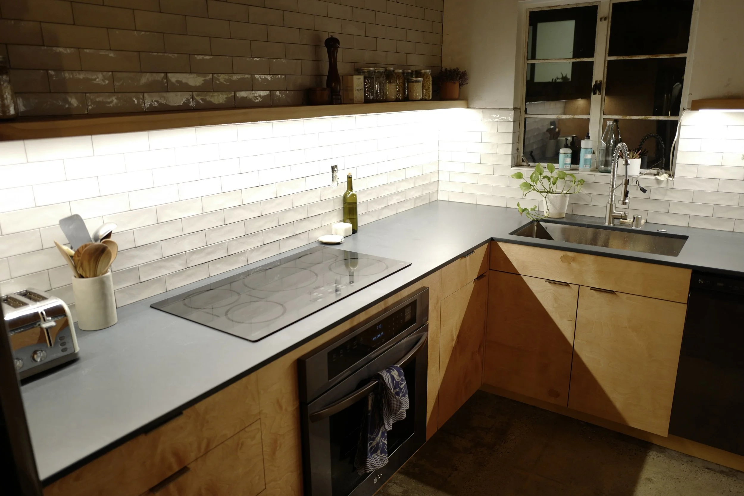 A cozy kitchen with a gray countertop, white subway tile backsplash, wooden cabinets, and a black oven and stovetop. There is a toaster, utensil holder, and a bottle of olive oil on the counter. A window above the sink shows a nighttime scene outside