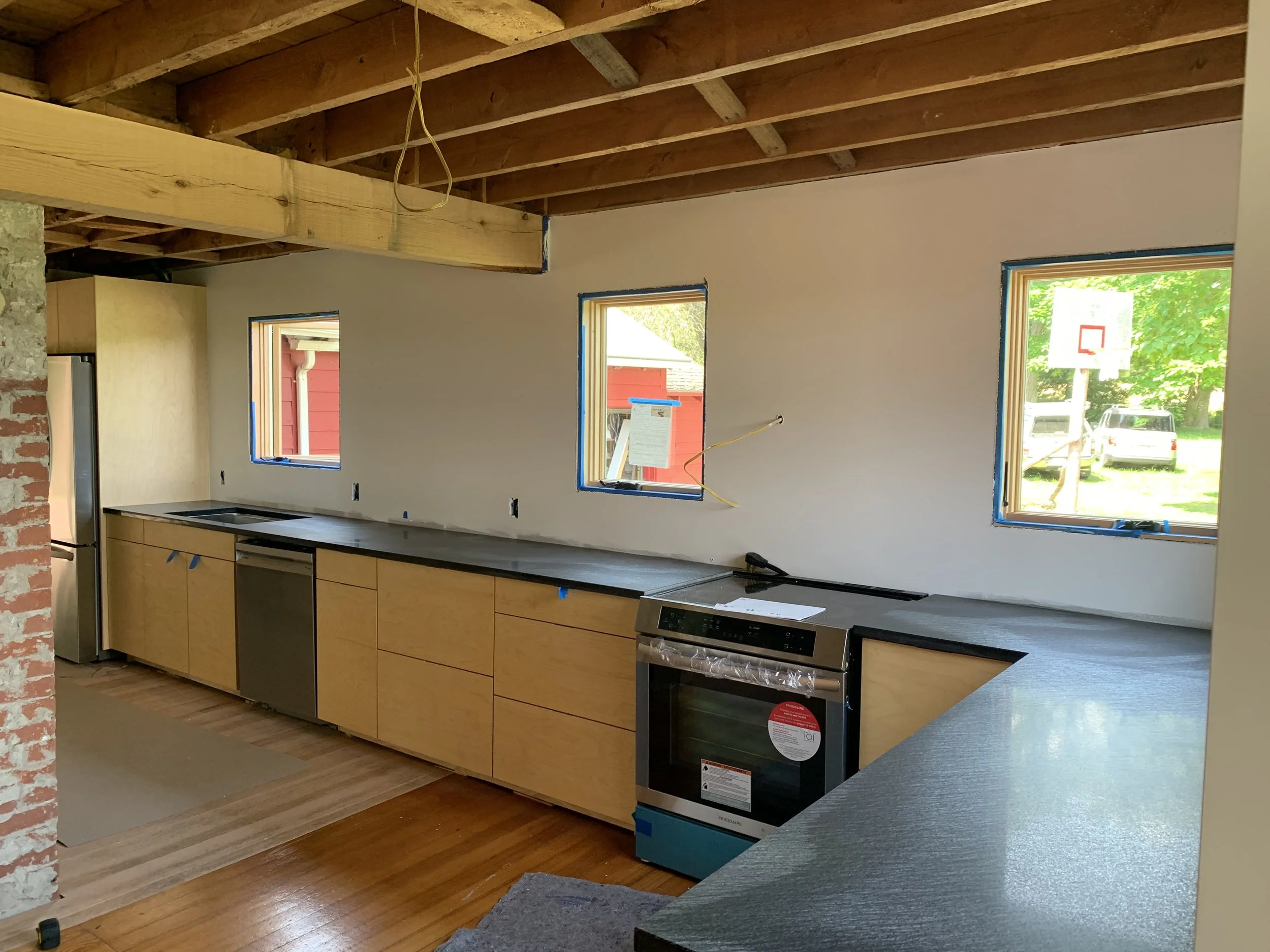 Kitchen under construction with new cabinets, stainless steel appliances, three windows, unfinished ceiling, and construction materials.