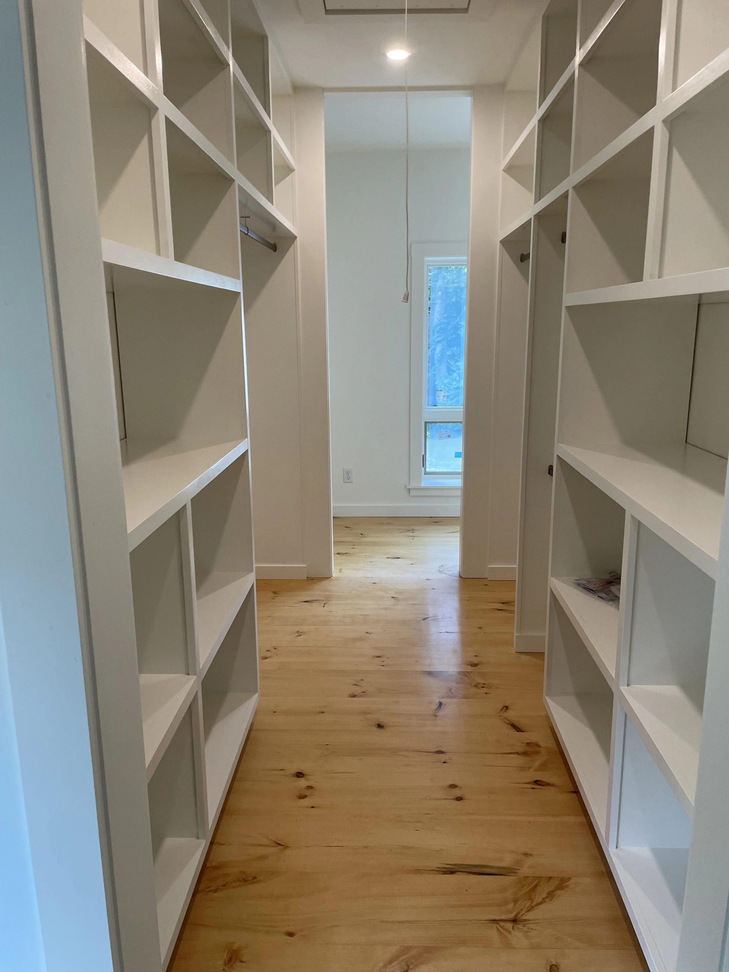 Empty walk-in closet with white built-in shelves on both sides, wooden floor, and a window at the end of the room with natural light.