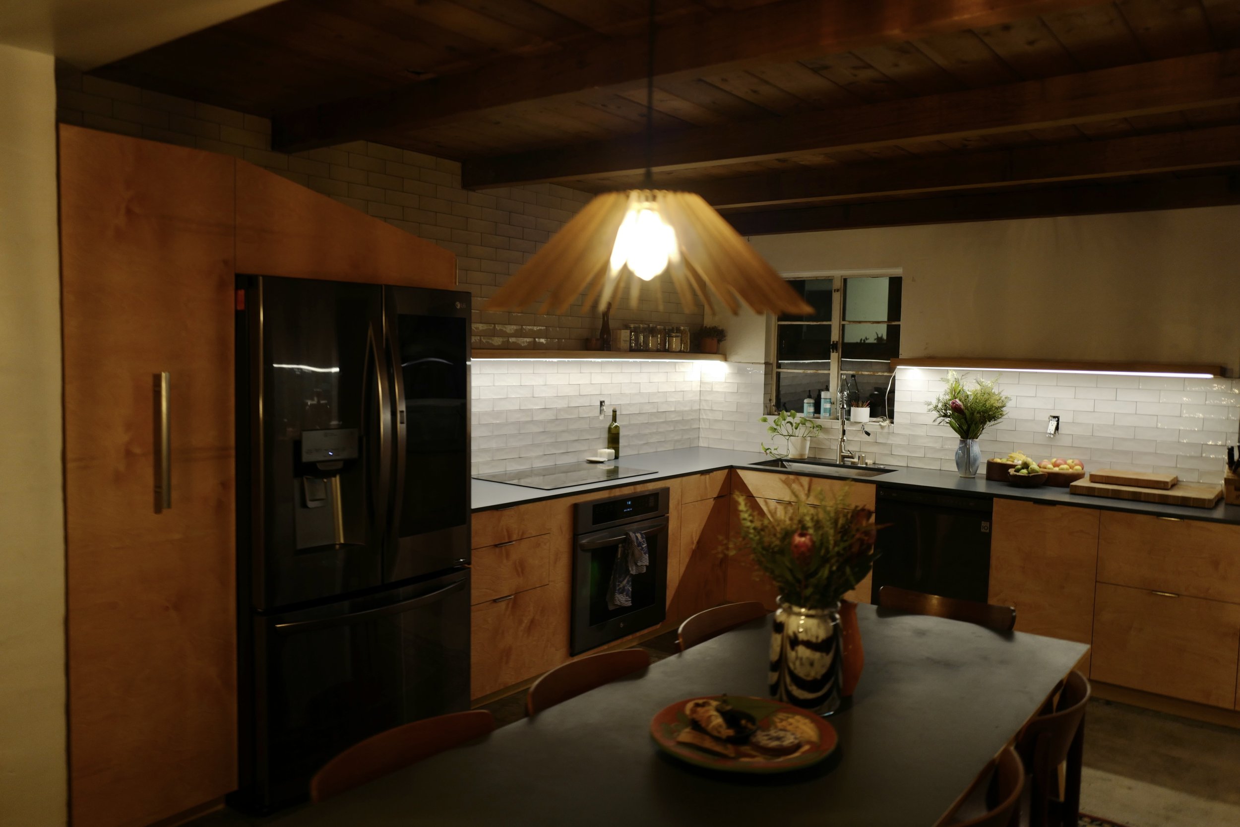 Kitchen with wooden cabinets, stainless steel refrigerator, oven, and white subway tile backsplash, illuminated by a pendant light, with a dining table and floral centerpiece.