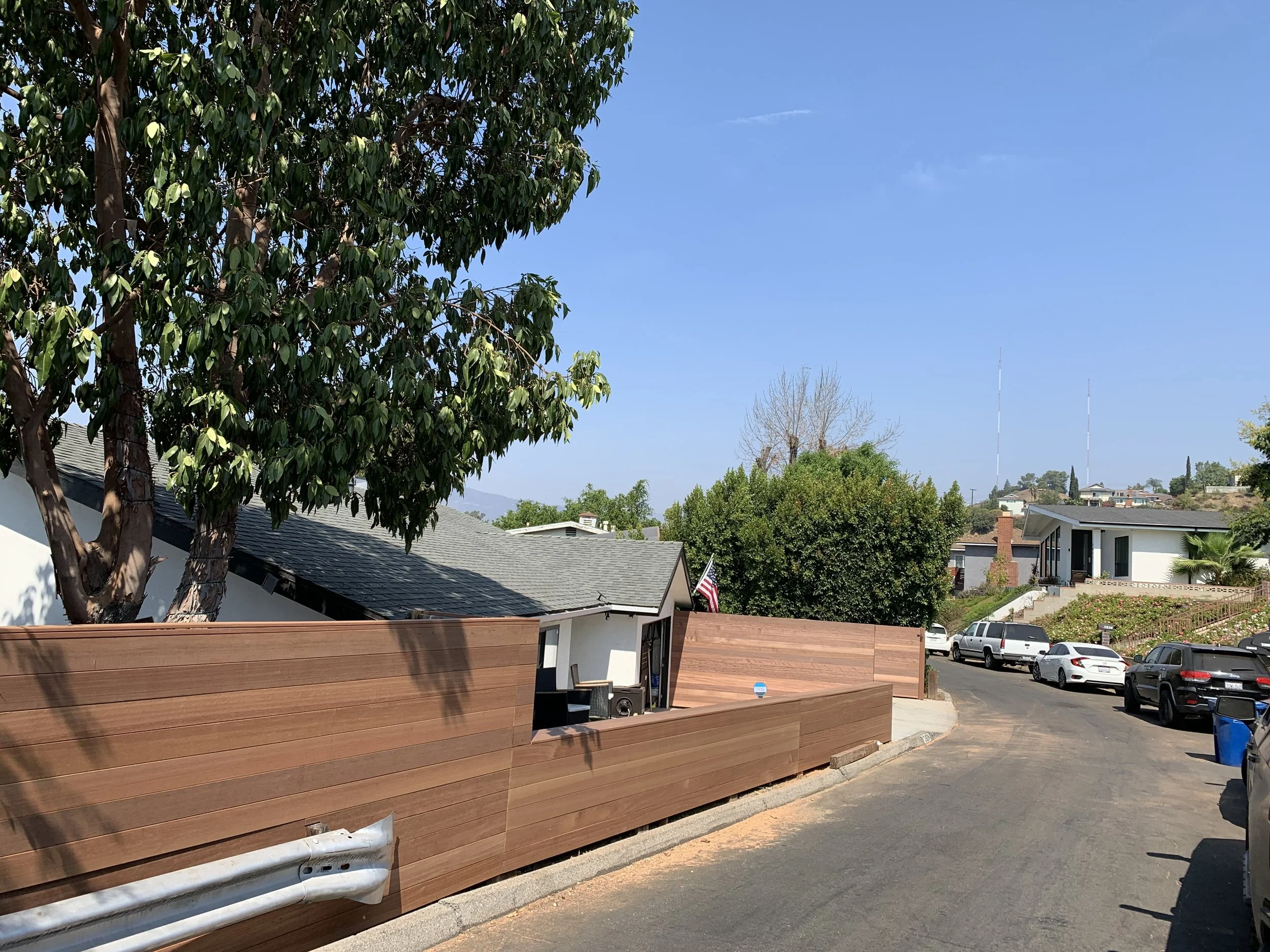 A residential street with houses, parked cars, and a large tree with green leaves. A wooden fence lines the sidewalk, and the sky is clear and blue.