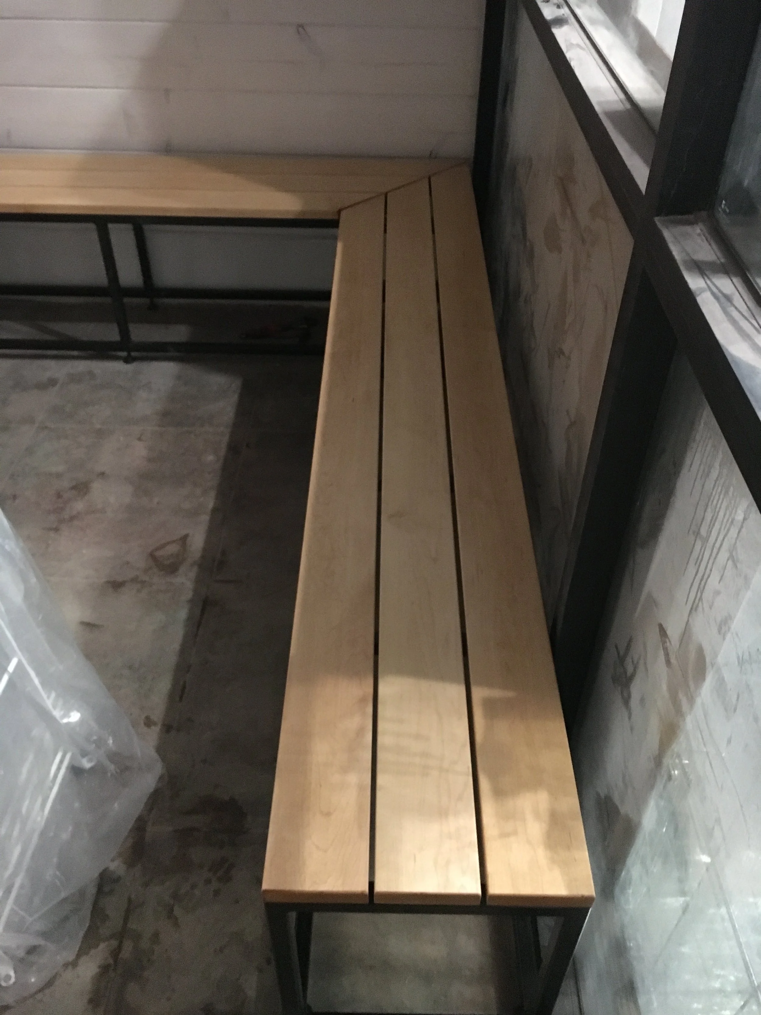 A wooden bench with three wooden planks on top, situated in a room with unfinished concrete flooring, a wood-paneled wall, and a window with a black frame.