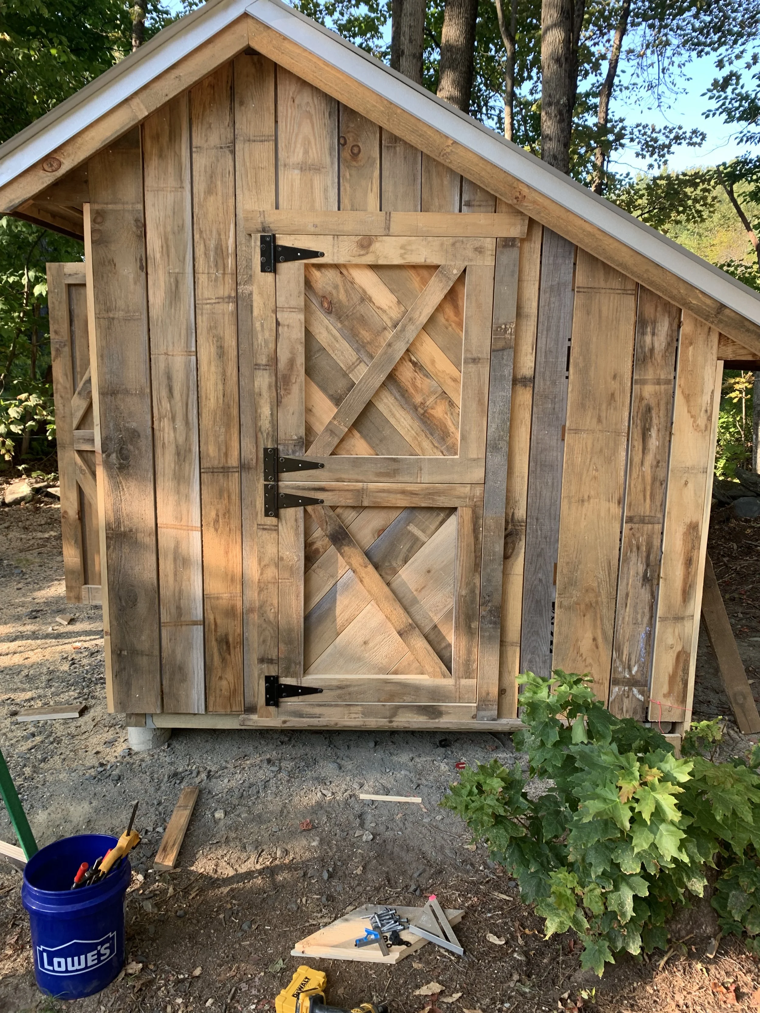 Small wooden shed with slanted roof, double doors with black hinges, surrounded by trees and a grassy area, construction tools and materials on the ground.