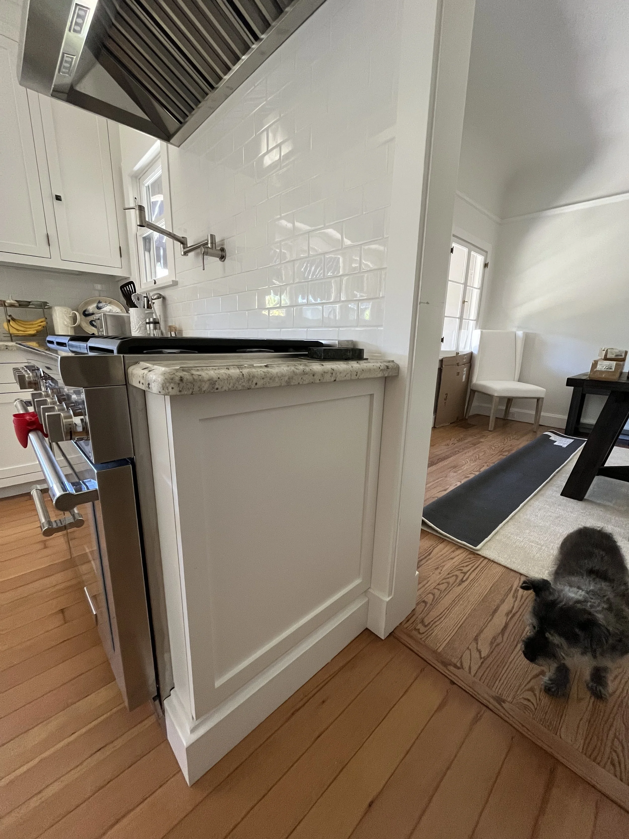 View of a kitchen with white cabinets, a white subway tile backsplash, a granite counter, and stainless steel appliances, including a stove and range hood, with a small dog on a wooden floor near the kitchen entry.