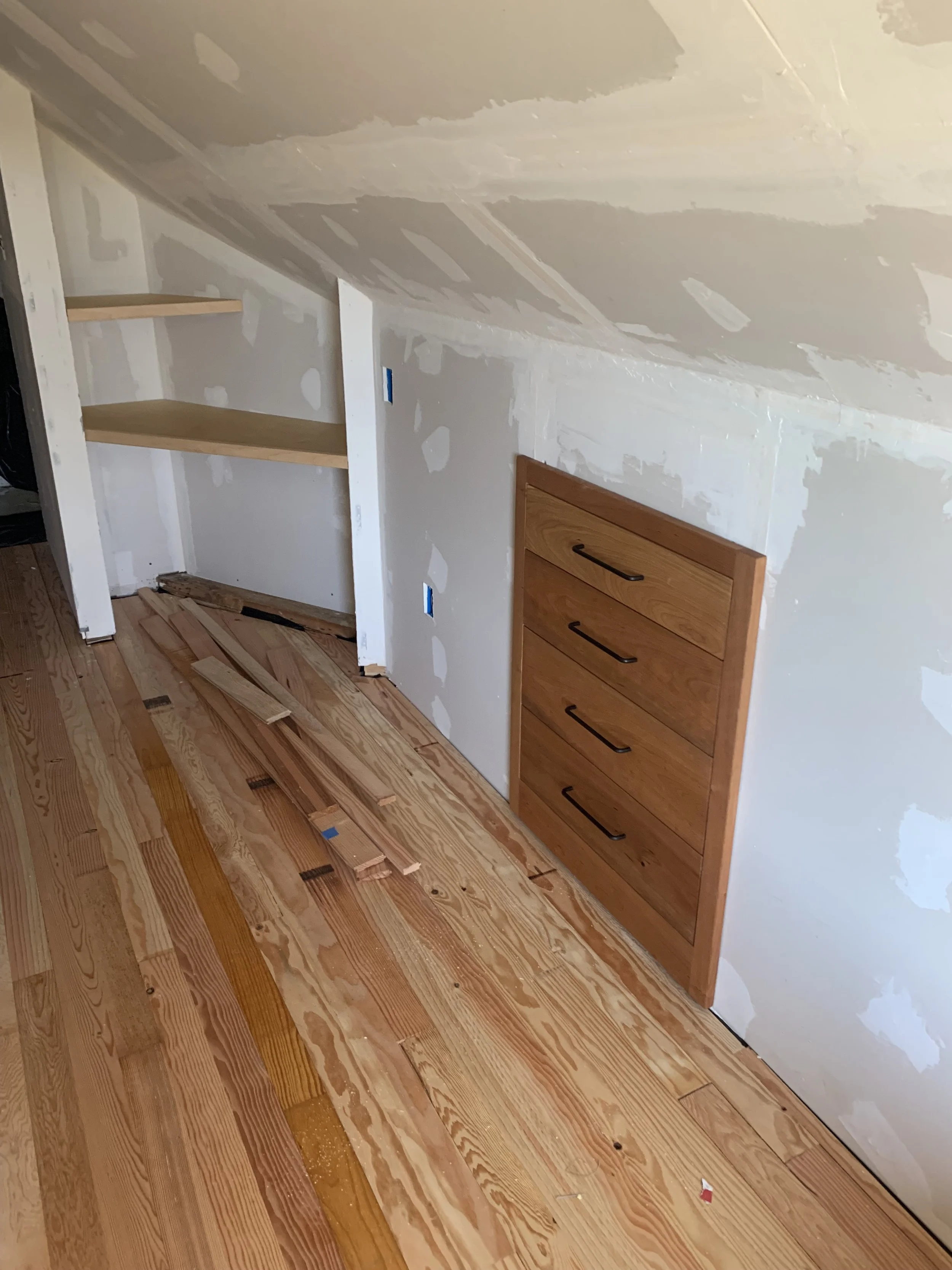 Interior of a room under construction with unfinished drywall on the walls and ceiling, a built-in wooden drawer unit, and hardwood flooring partially installed with some loose and cut wooden planks on the floor.