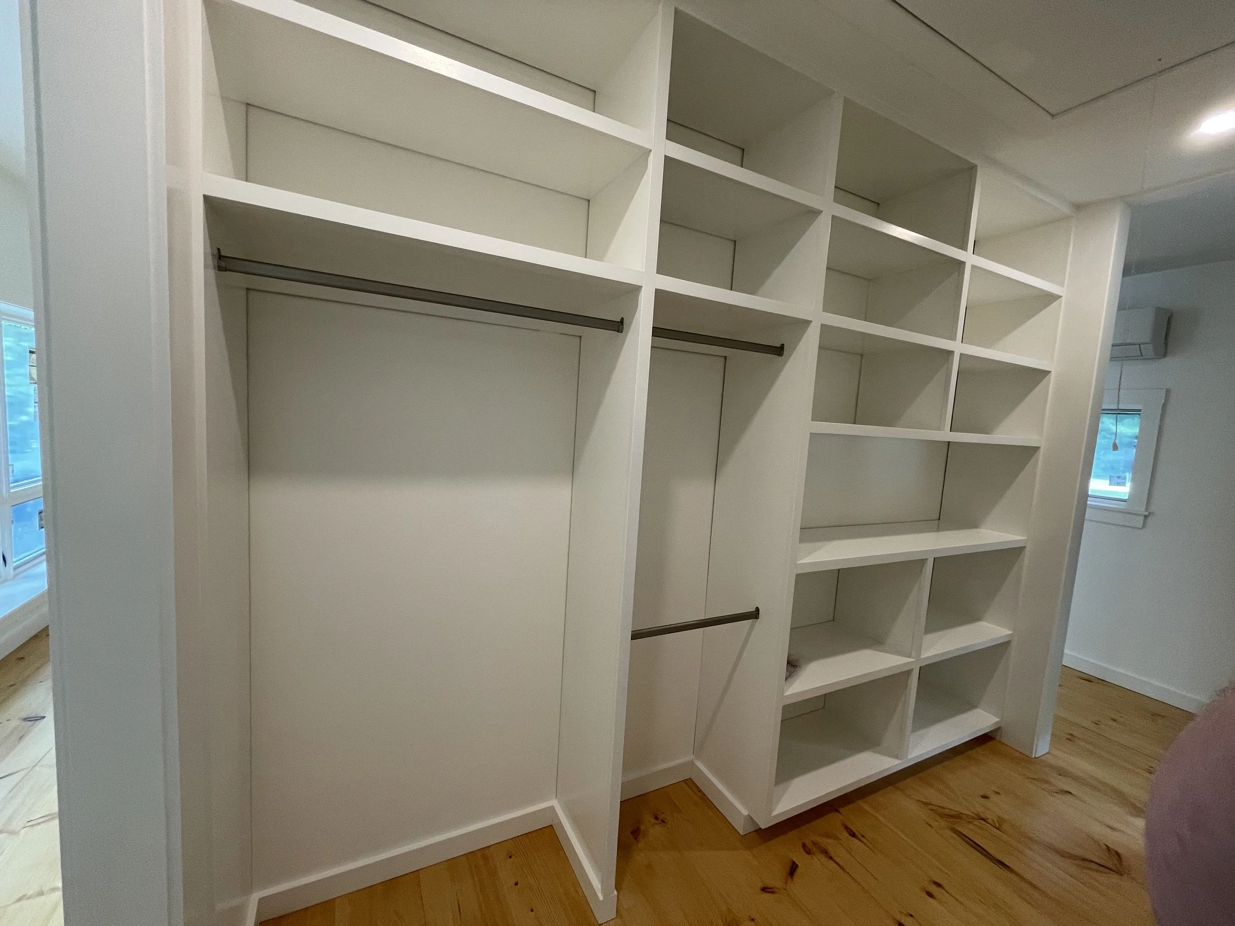 Empty white built-in closet with multiple shelves and hanging rods, wood floor, and two windows in a room.