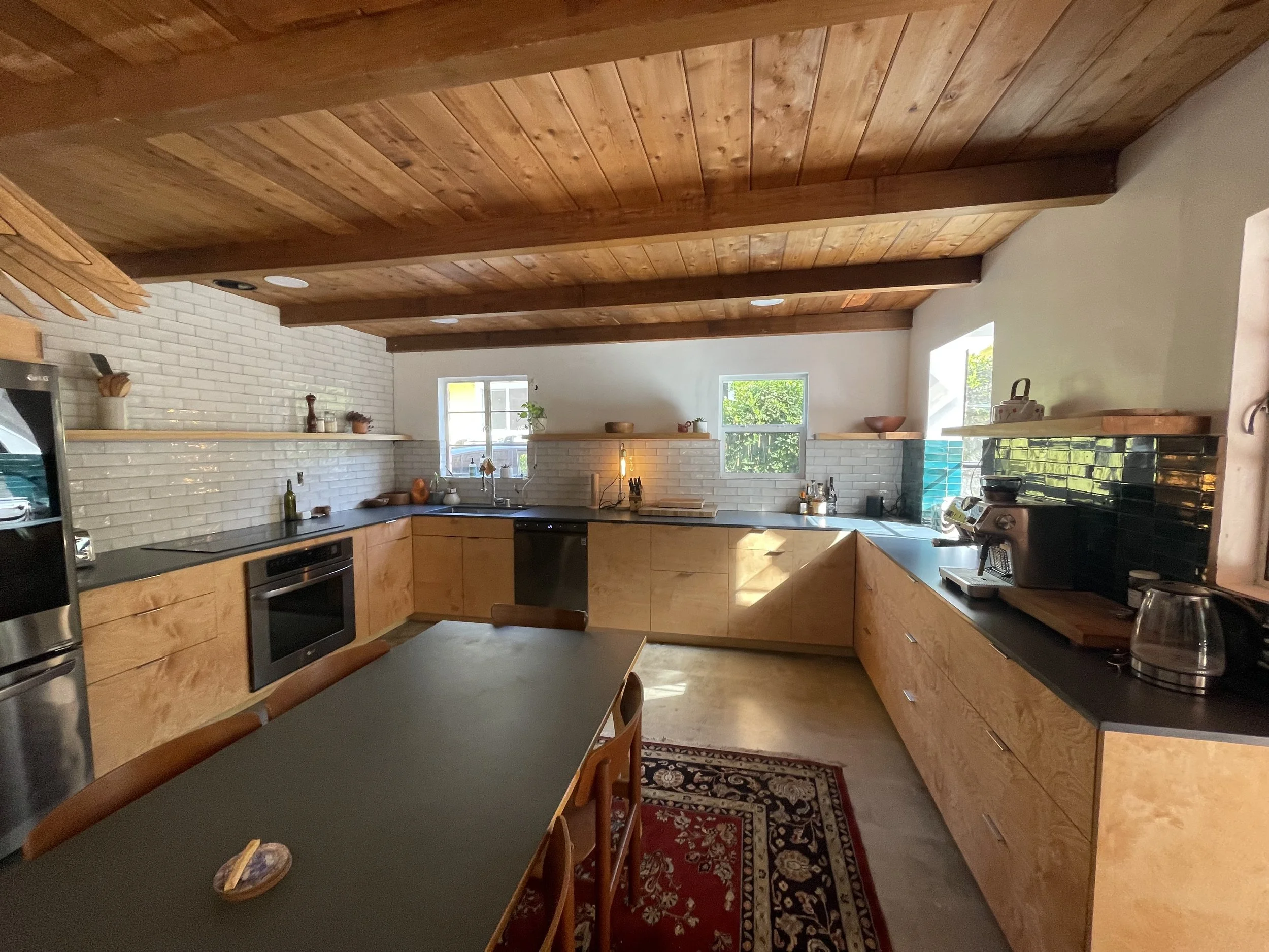 Kitchen with wooden cabinets, black countertops, white brick backsplash, and wooden ceiling. There are three windows, a dining table with chairs, and various kitchen appliances and decorations.