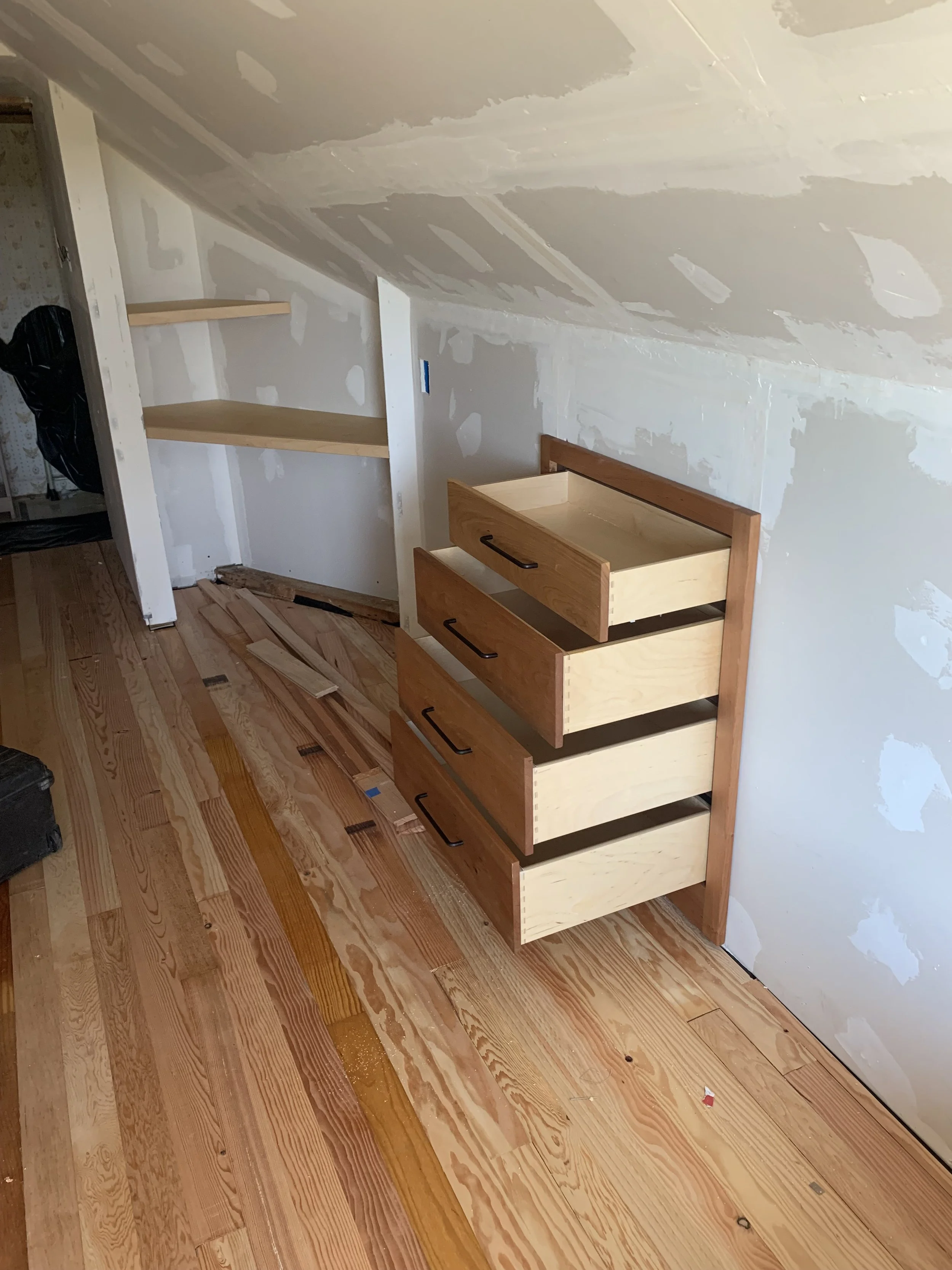 Wooden dresser with five open drawers in a room under construction, unfinished walls, and a sloped ceiling.