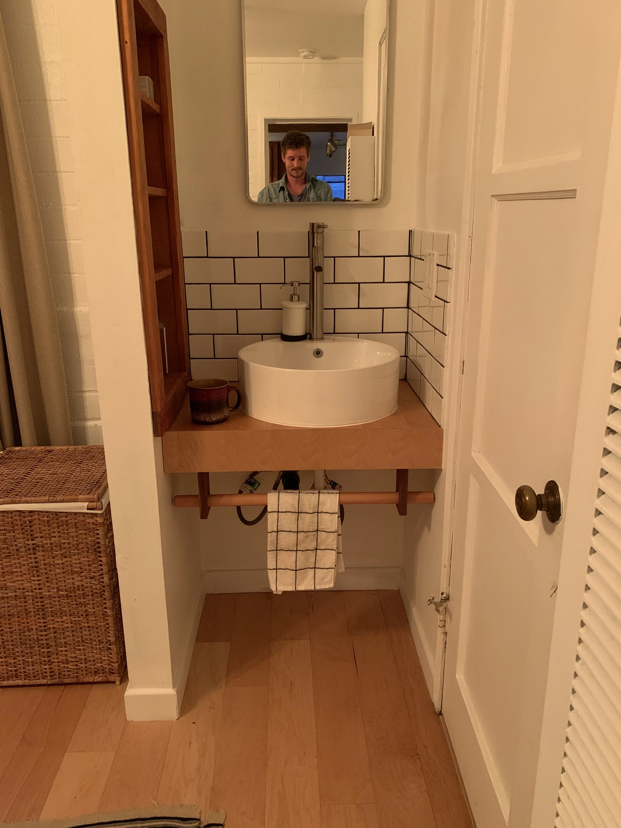 A small bathroom with a round white sink on a wooden countertop, a mirror above the sink, and a person taking a selfie visible in the mirror. The wall behind the sink has white subway tiles, and there is a soap dispenser on the sink. To the left, the