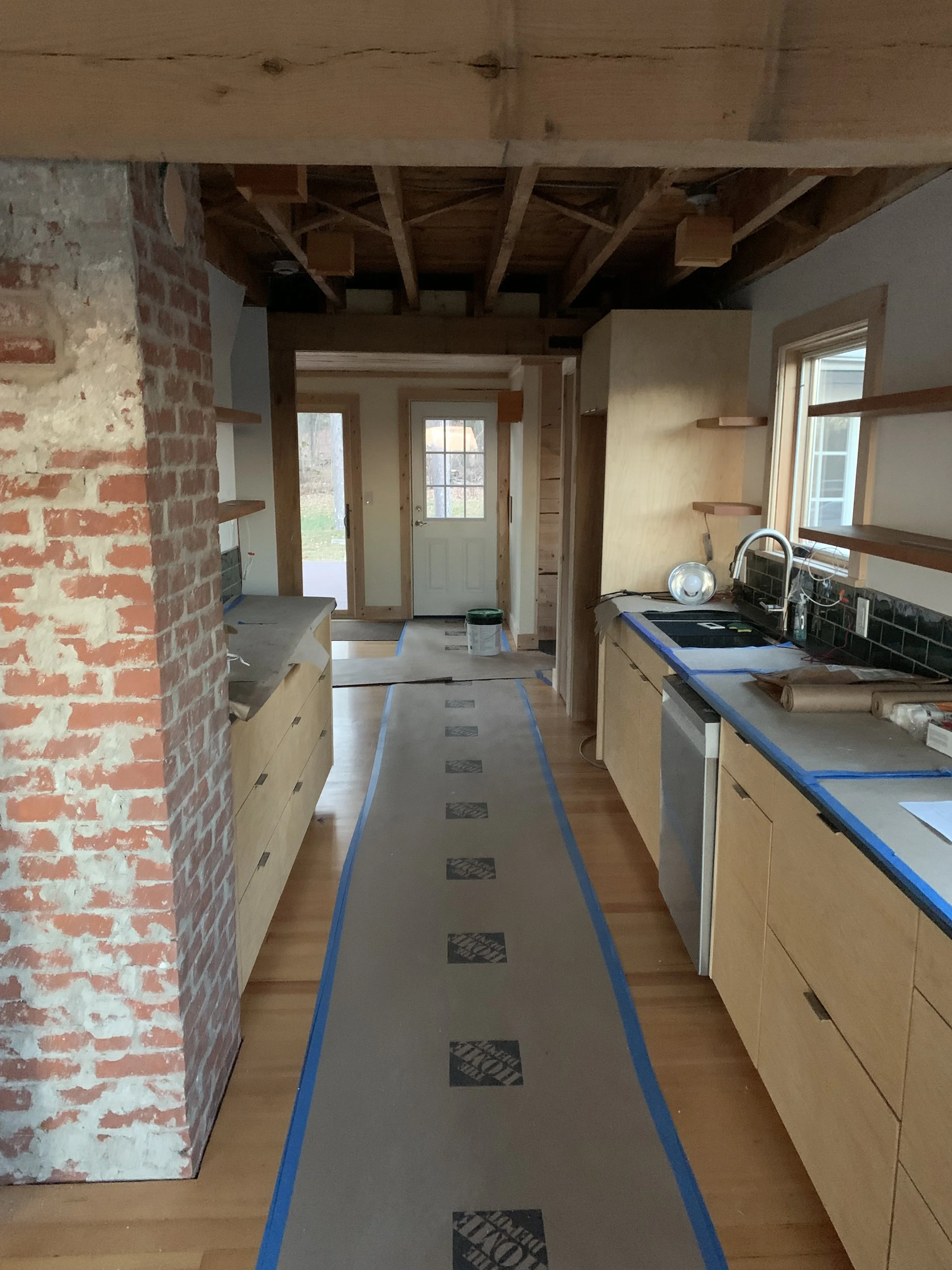 Interior of a kitchen under construction with wood cabinets, exposed brick wall, and plywood ceiling. Blue painter's tape and protective paper on the floor, with a door at the back leading outside.
