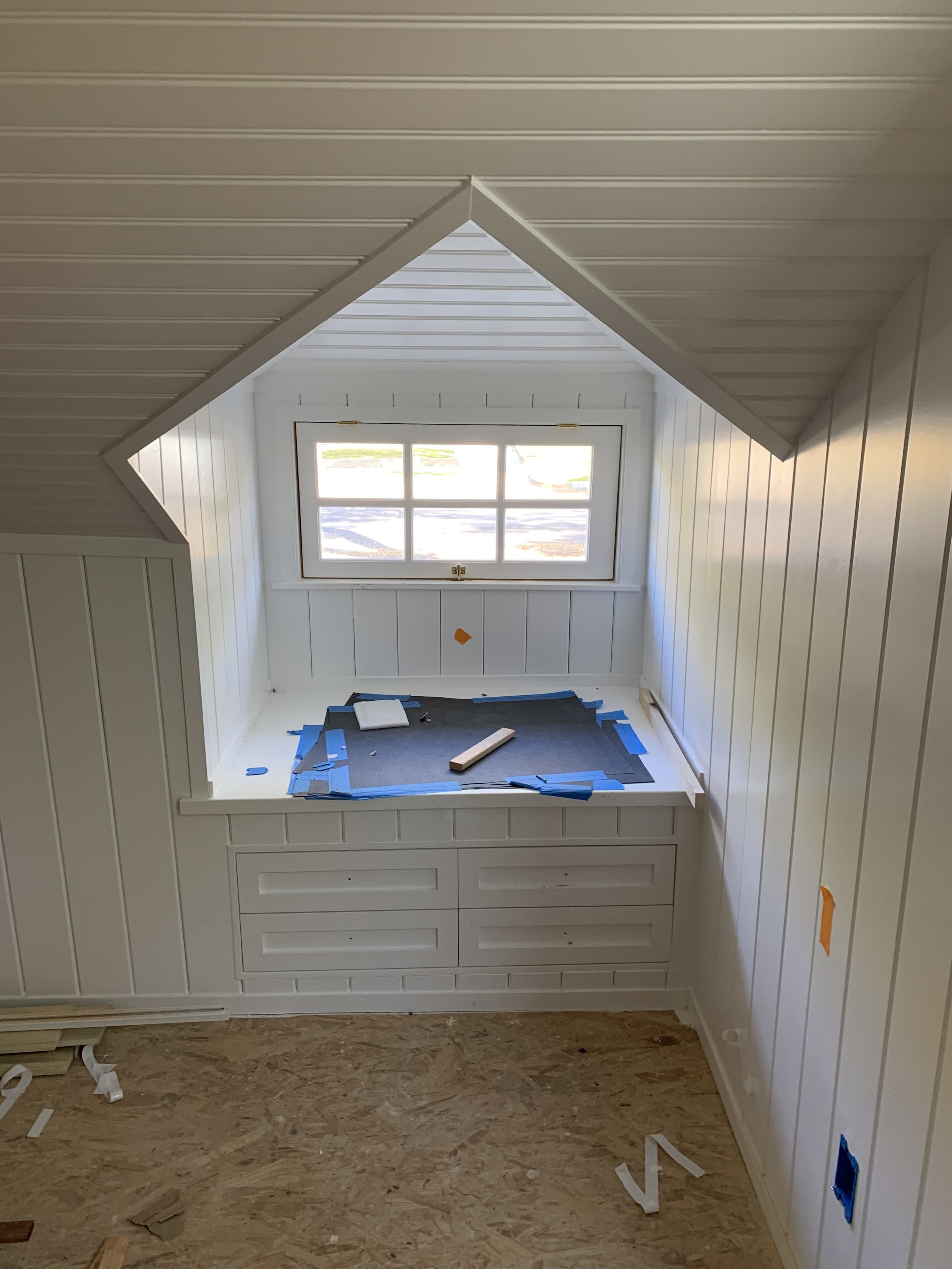 A room under construction with white paneled walls, a sloped ceiling, and a window. There is a built-in window seat covered with blue painter's tape and construction materials on the floor.