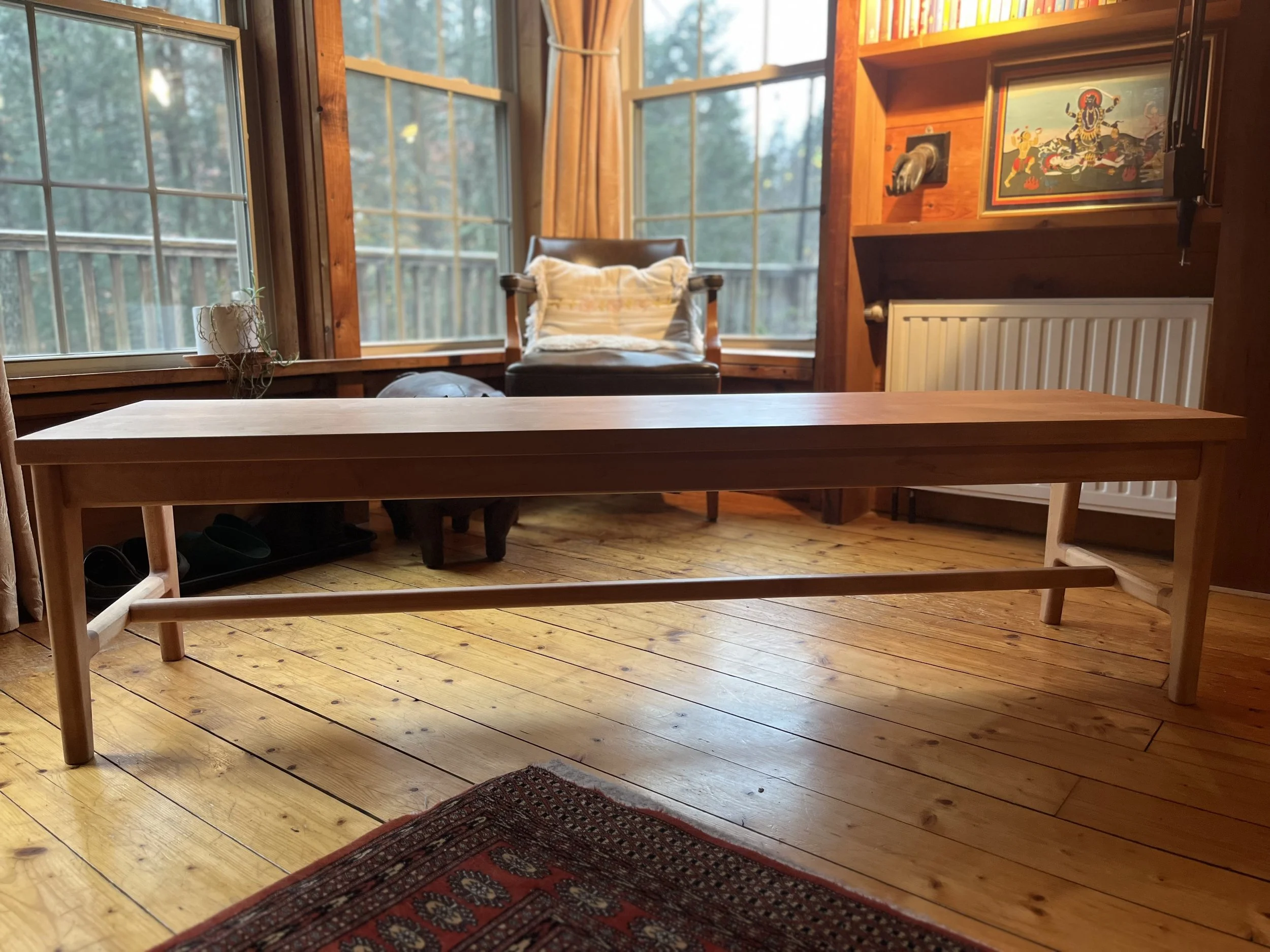 A wooden bench indoors in front of large windows with a view of trees outside. A chair with a pillow and cushions is in the corner, a small plant on the windowsill, and artwork on the wall. The flooring is wooden, and a colorful rug is partially visi