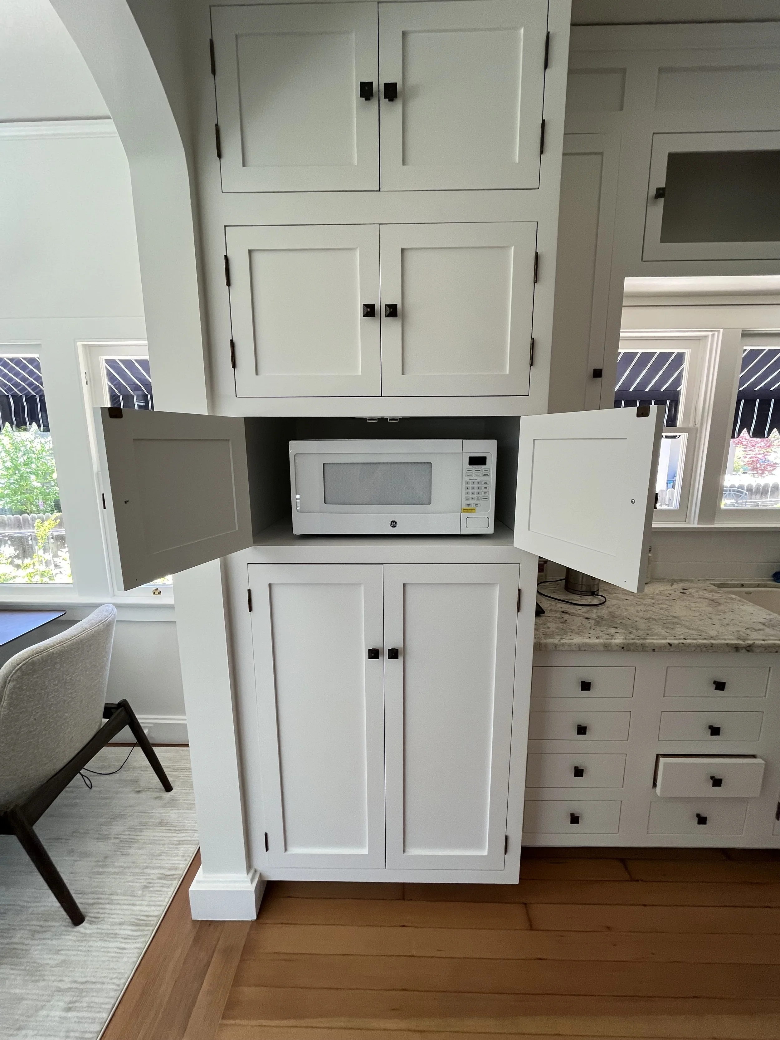 Kitchen cabinet with two open doors showing a microwave inside, and multiple closed cabinets above and below, with a granite countertop to the right and windows in the background.