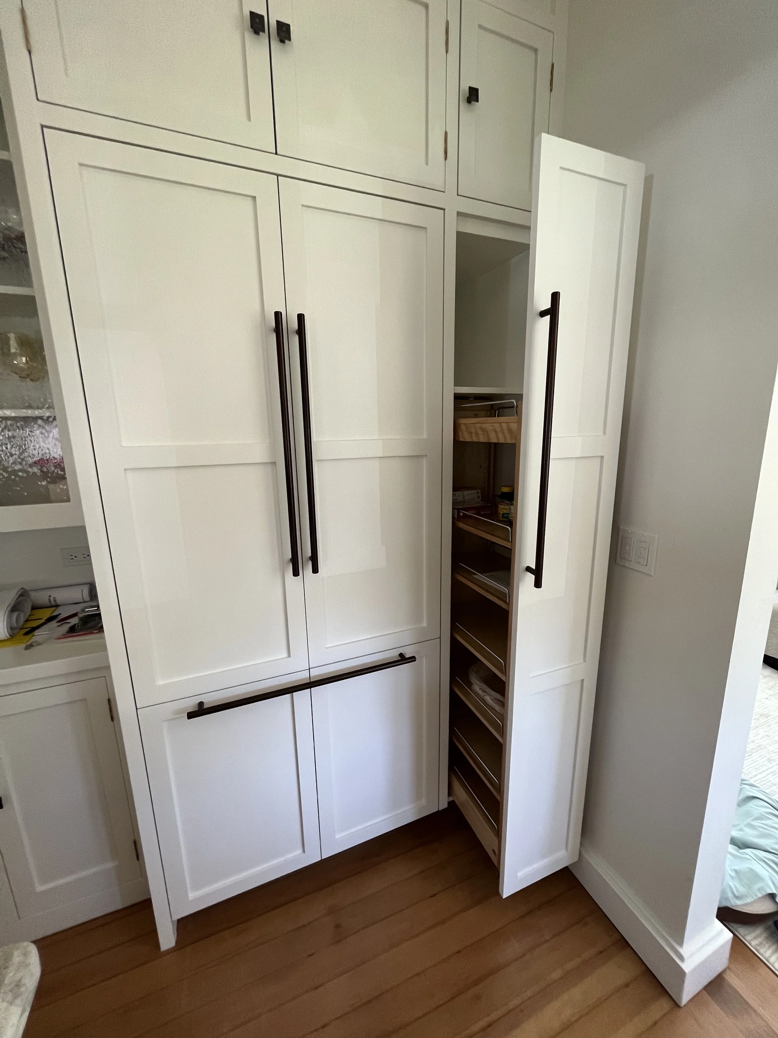 A tall white kitchen cabinet with black handles, partially open revealing shelves inside, next to a white wall and on a wooden floor.