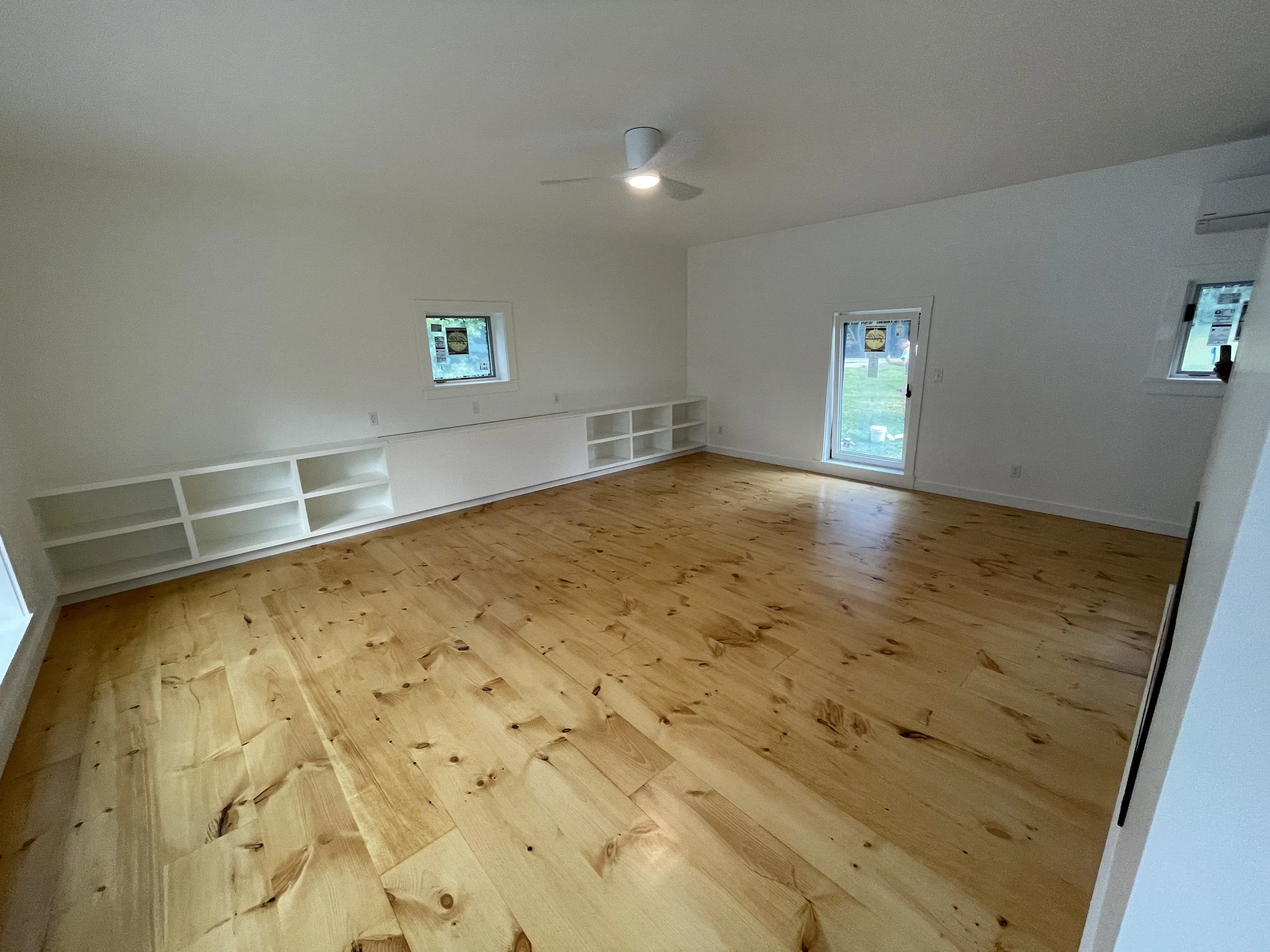 Empty room with white walls, hardwood floor, three windows, a white built-in shelf, and a ceiling fan with light.