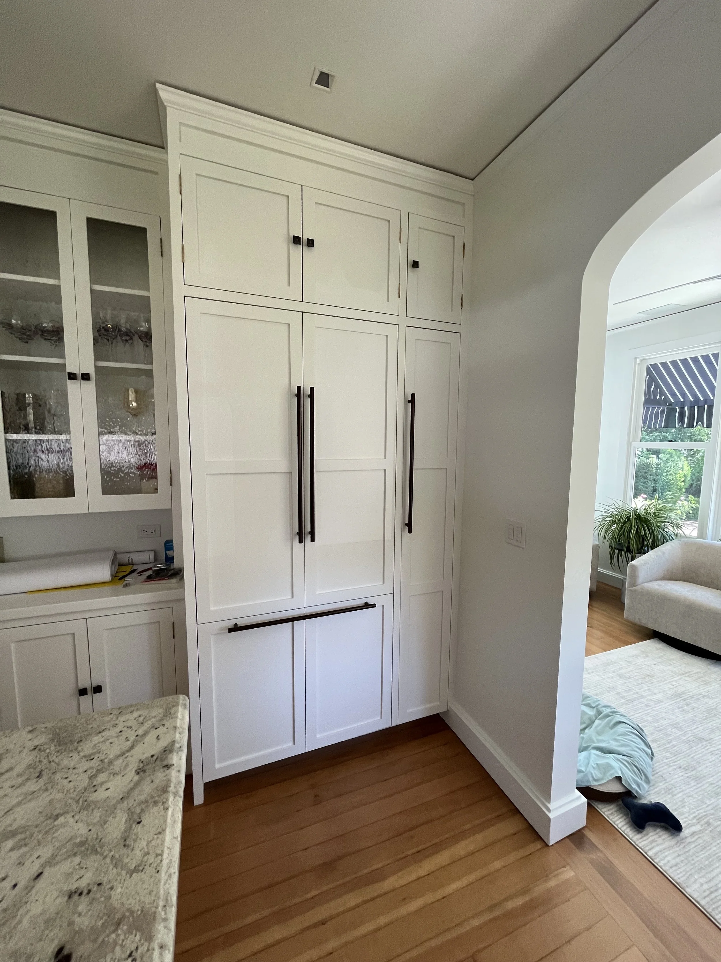 White built-in cabinets in a kitchen, with black handles and a view into a living room with a couch and a window with a striped awning.