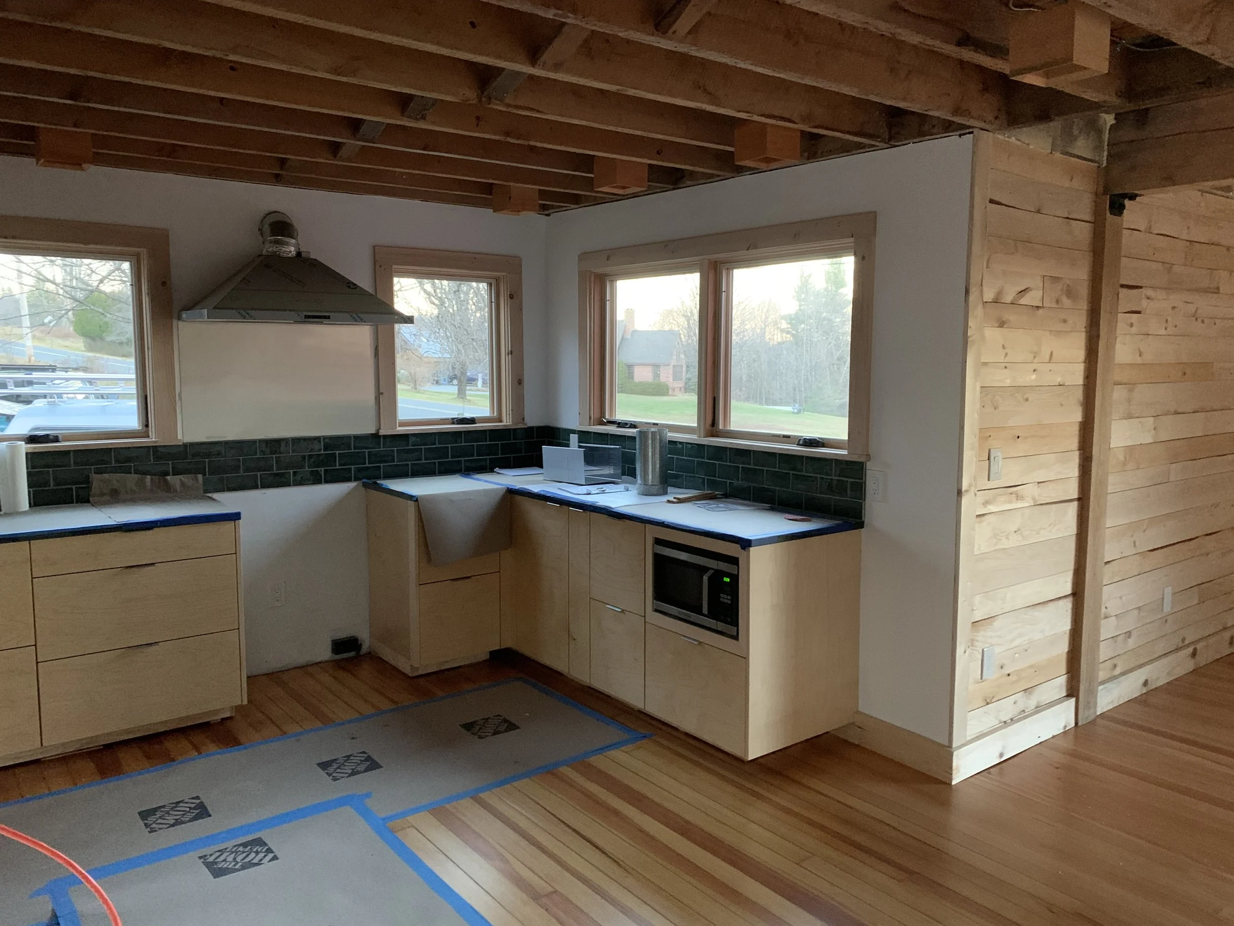 Kitchen under construction with light wood cabinets, black subway tile backsplash, a stainless steel range hood, large windows, and wooden ceiling beams.