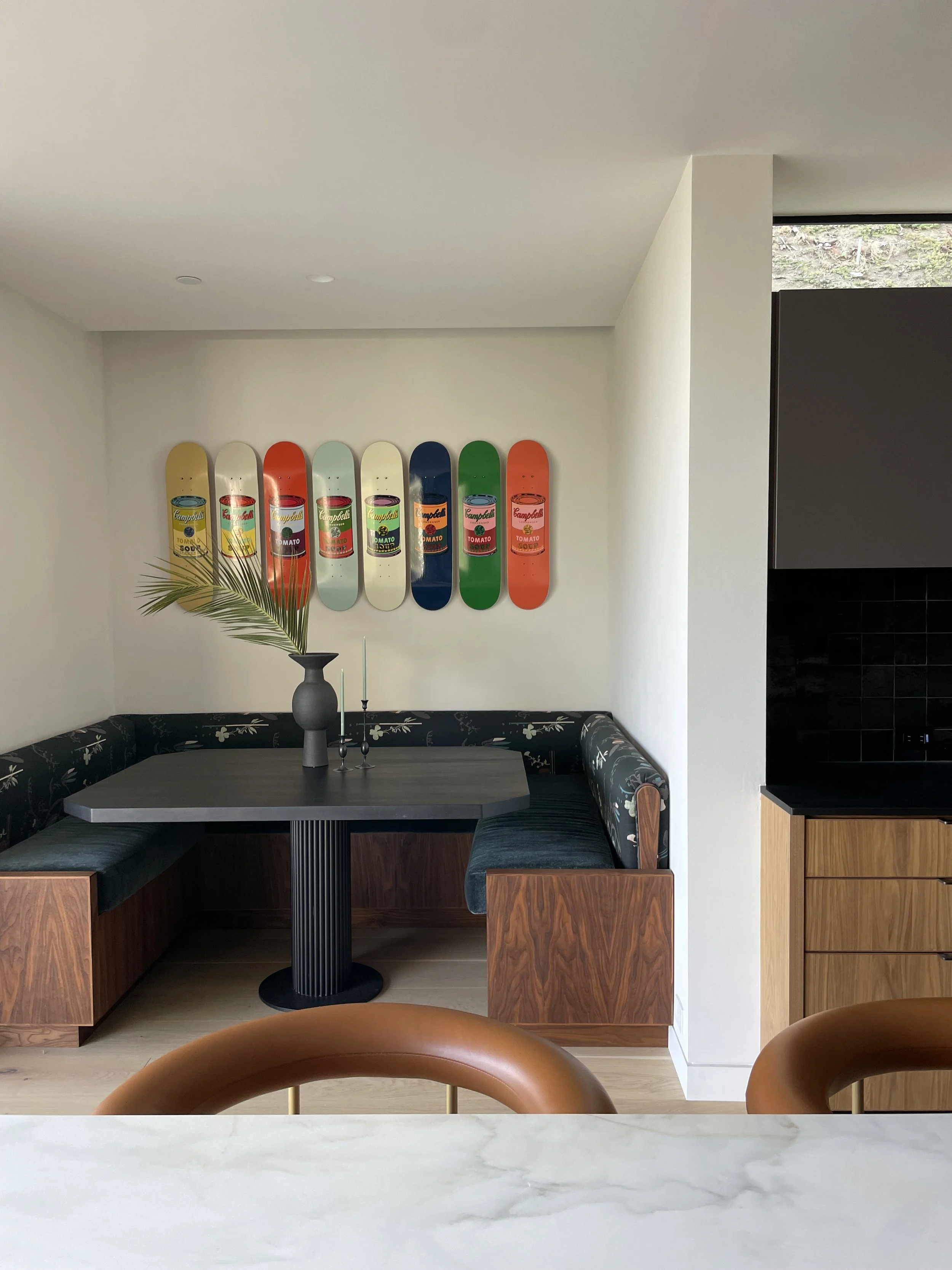 Interior of a modern dining area featuring a built-in corner booth with dark green cushions and a dark gray table. On the wall behind the booth, there are seven skateboard decks decorated with colorful pop art resembling Campbell's soup cans, mounted