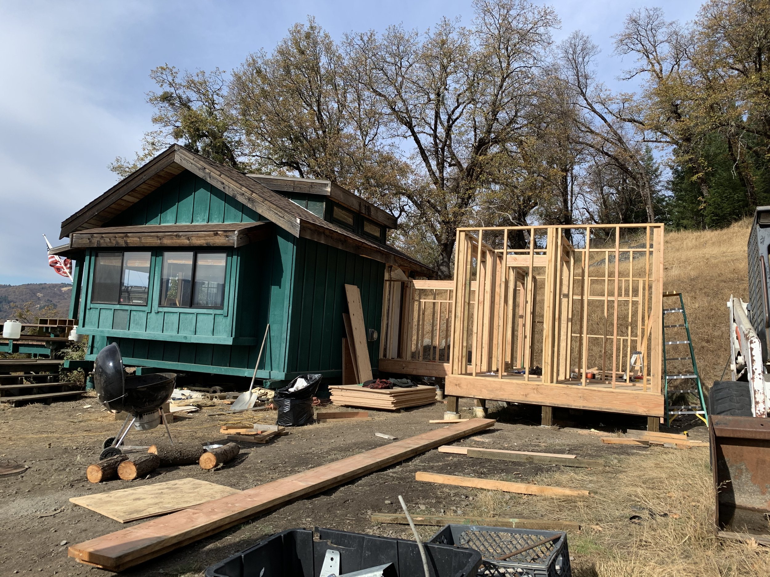 Construction site with a small teal house and a wooden frame structure beside it, surrounded by tools and building materials, with leafless trees in the background.