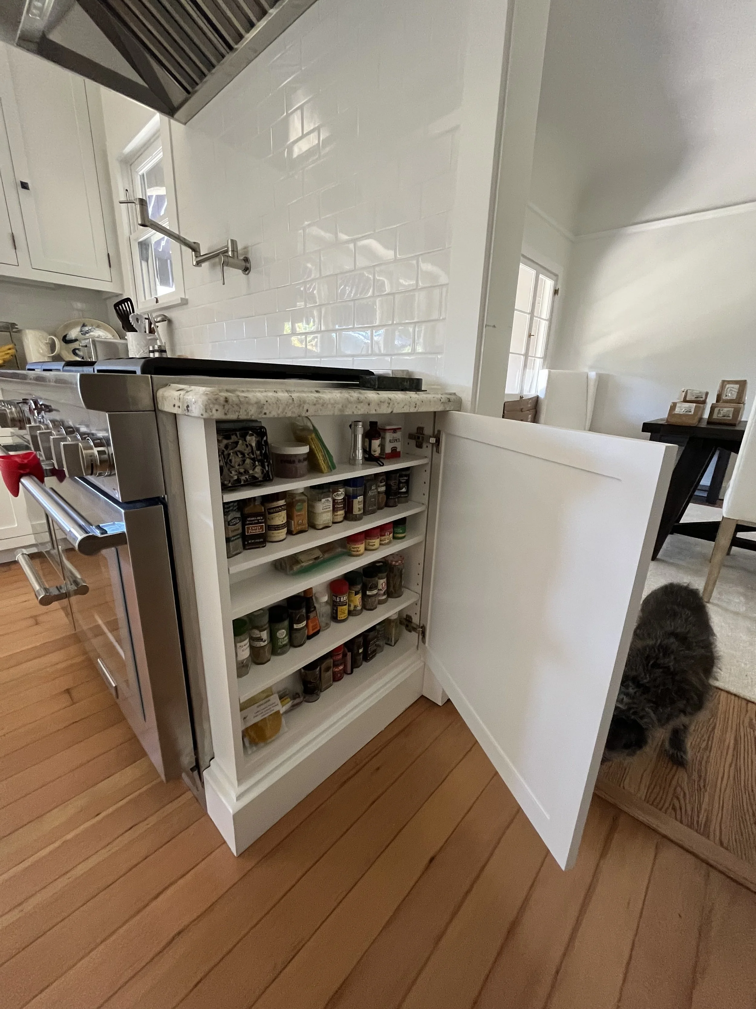 Open kitchen cabinet filled with various spices, located next to a stove with a marble countertop, in a home kitchen with hardwood floors and white cabinetry.